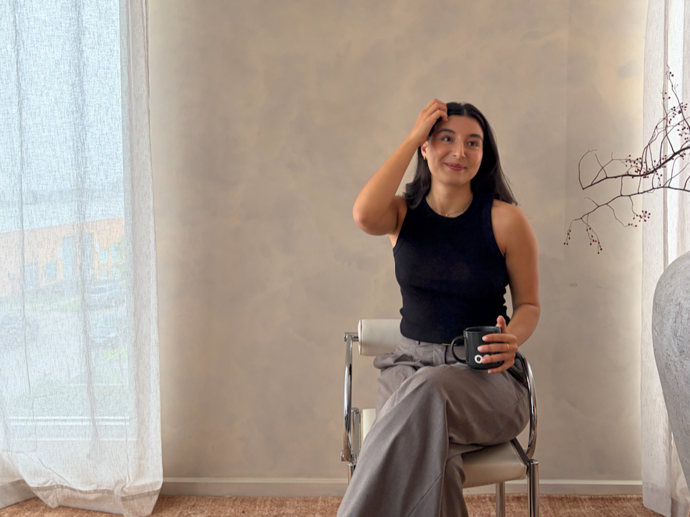 A woman sitting on a chair indoors, holding a black mug, with her right hand touching her forehead, and smiling.
