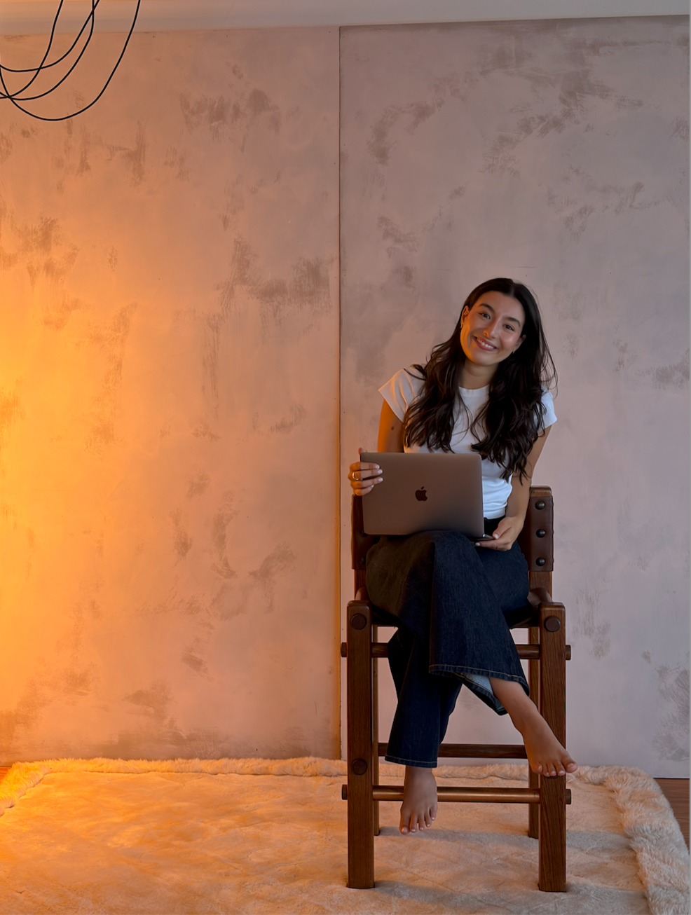 A woman with long dark hair sitting on a wooden chair, smiling and holding a silver MacBook laptop, indoors with warm lighting on a soft beige rug.