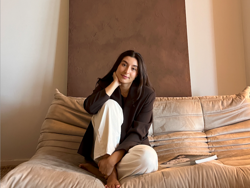A (naturopath) woman sitting cross-legged on a beige sofa, resting her chin on her hand, wearing a black blazer and white pants, with a relaxed expression in a room with a textured wall behind her.