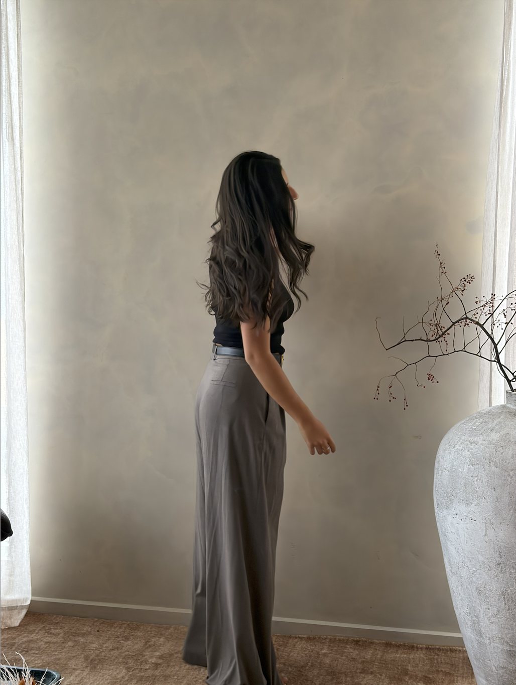 Side view of a woman with long, wavy dark hair, wearing a black top and gray wide-leg pants, standing indoors near a large vase with decorative branches.