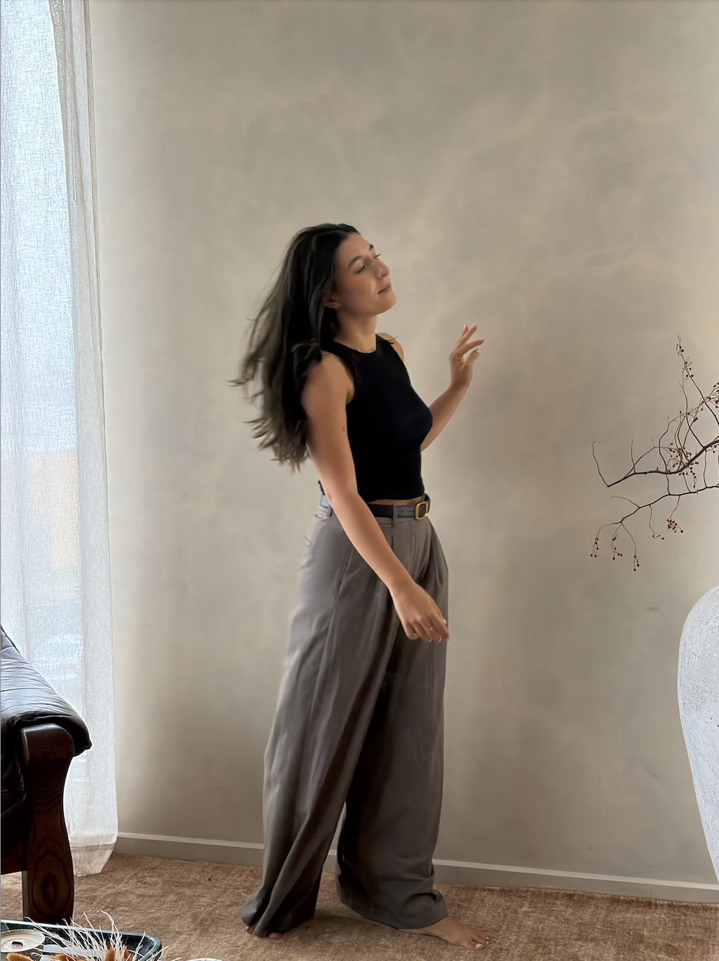 A woman standing barefoot in a room with beige walls and a textured wall, wearing a black sleeveless top and high-waisted gray pants, with her eyes closed and hand raised softly.