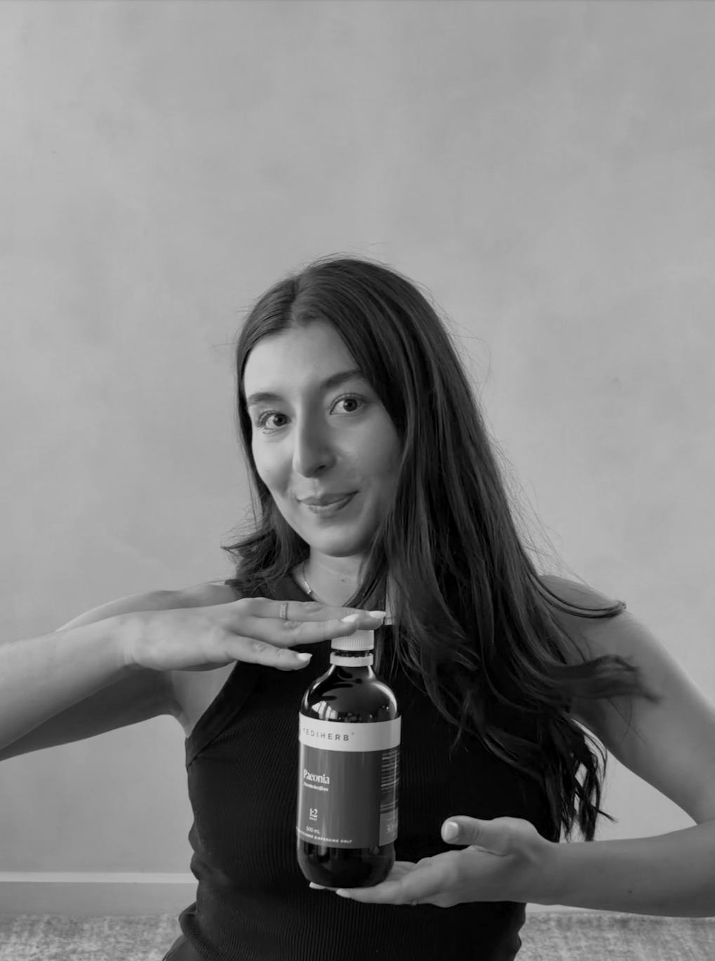 A woman with long dark hair holding a herbal MediHerb bottle labeled 'Peonia' in front of her chest, posing for a photo in front of a plain background.