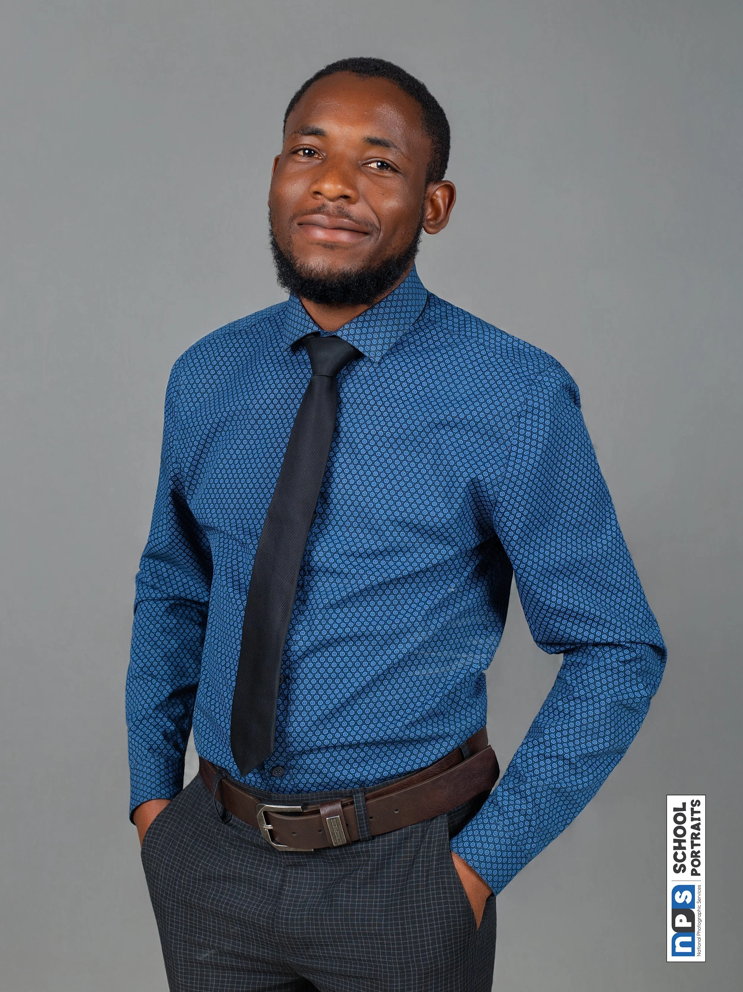 A young man with dark skin, short hair, and a beard leaning against a beige wall outdoors during daytime. He is wearing a white polo shirt and black trousers, with a relaxed expression, in a casual pose with one hand hanging down and the other in his pocket.