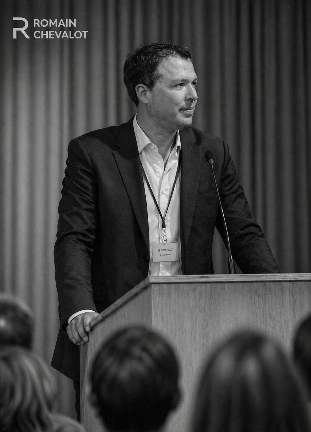 A man in a suit standing at a podium, speaking at a conference, with a microphone in front. There is a curtain behind him, and audience members are visible in the foreground.