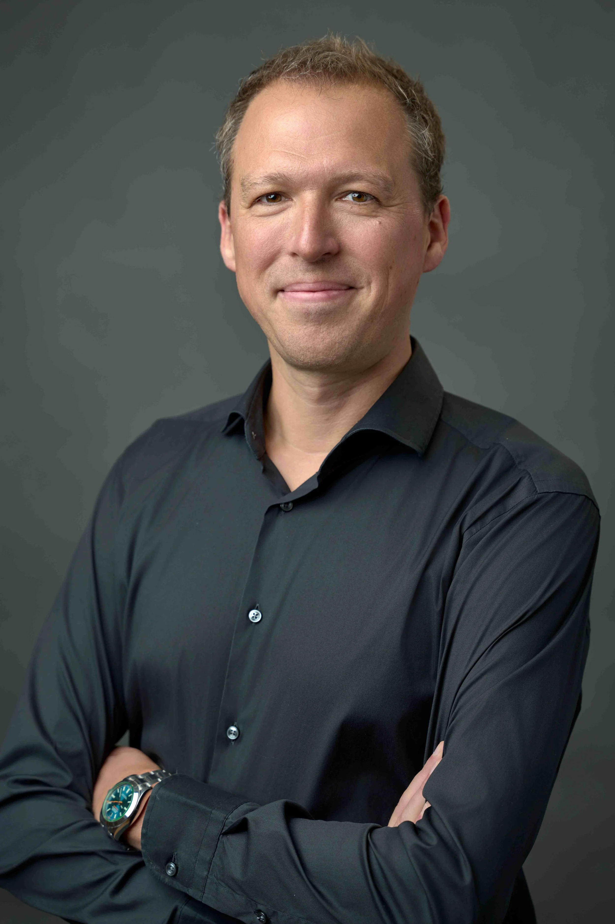 Professional man with light skin, short curly hair, wearing a black button-up shirt and a silver watch, smiling with crossed arms against a dark green background.