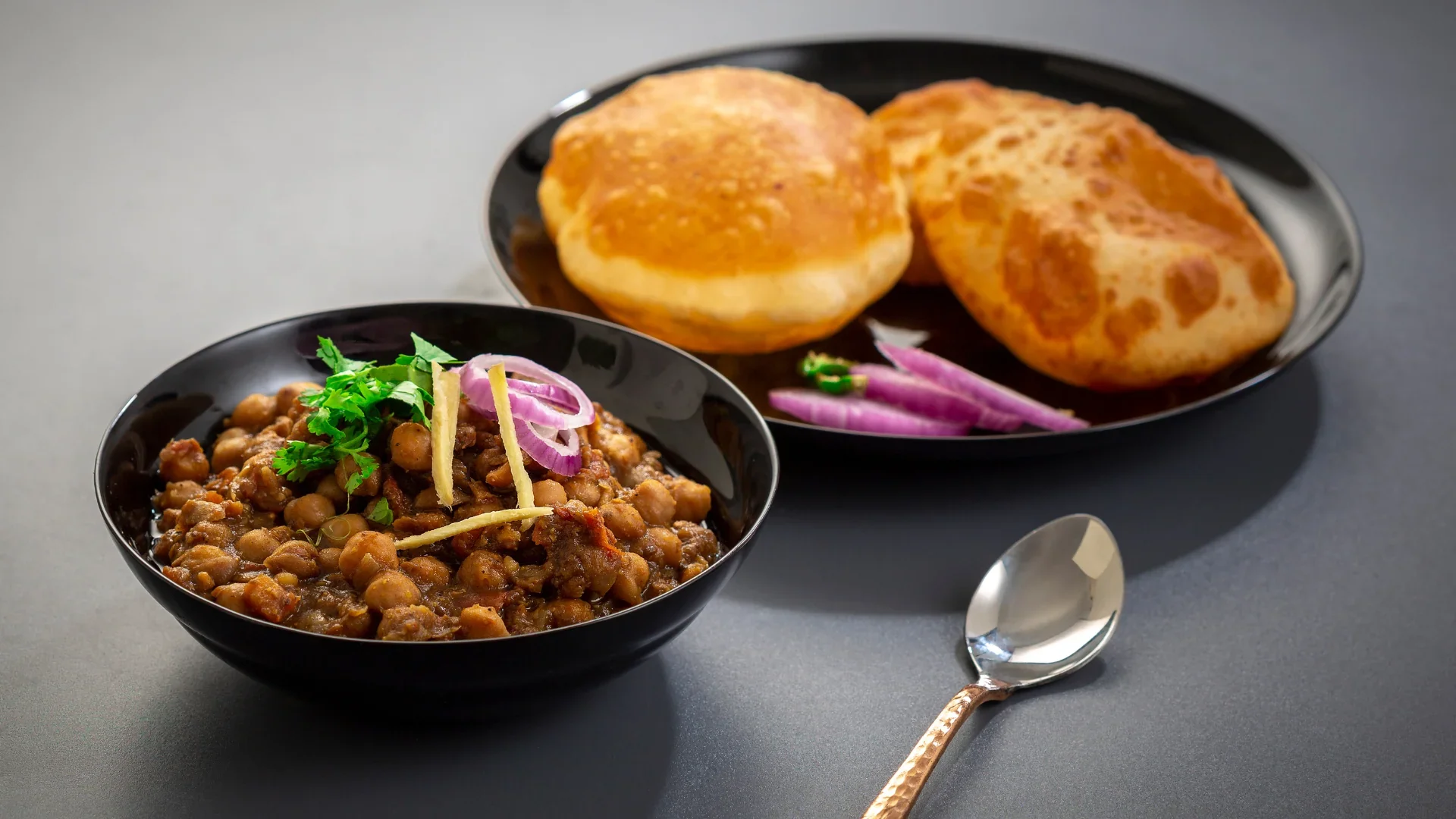 A black bowl of cooked lentils garnished with cilantro, sliced onions, and ginger pieces, placed in front of a black plate with two Indian bread rolls and sliced onions, on a gray surface.