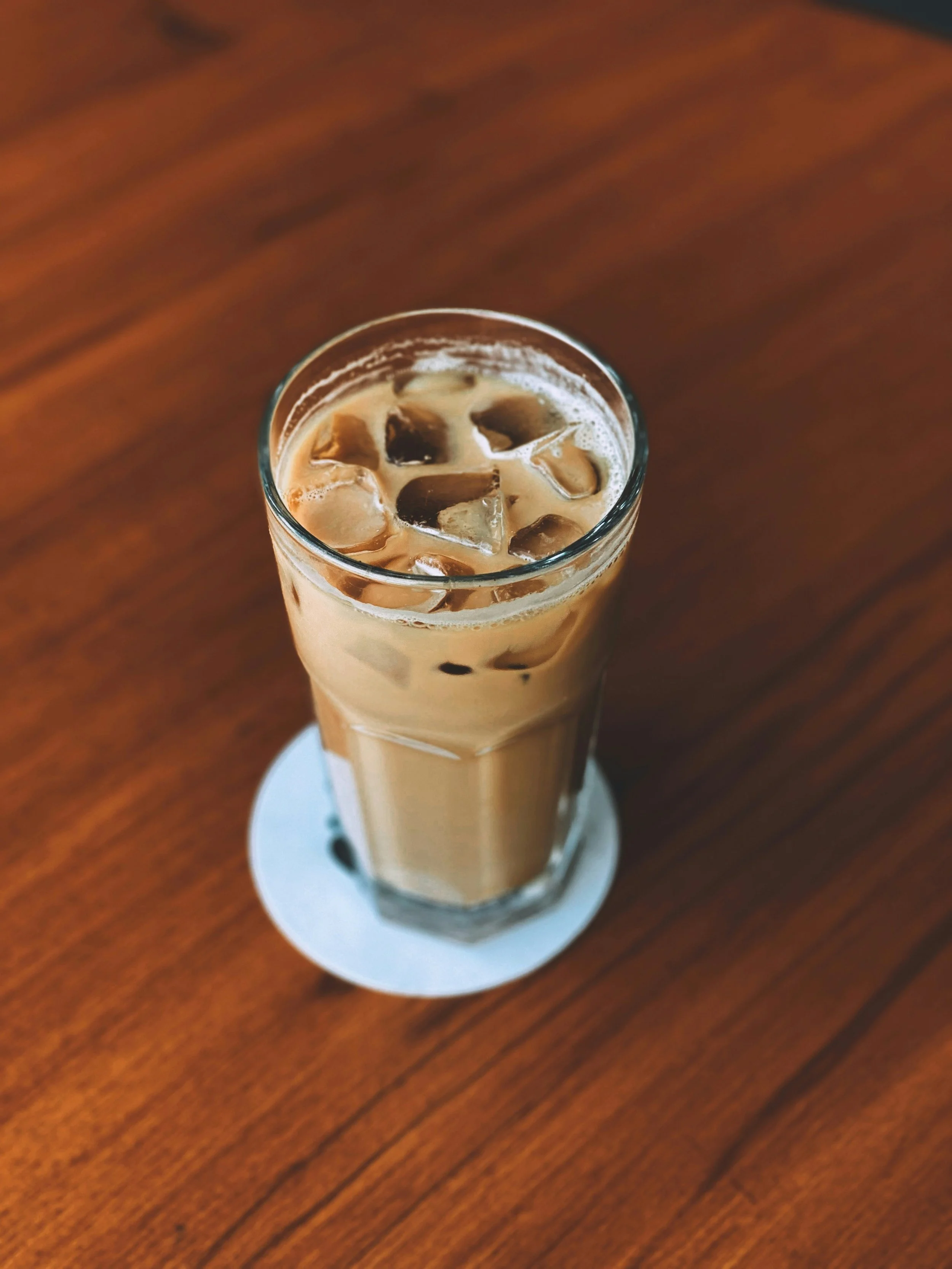 A tall glass of iced coffee with milk, filled with ice cubes, sitting on a white coaster on a wooden table.