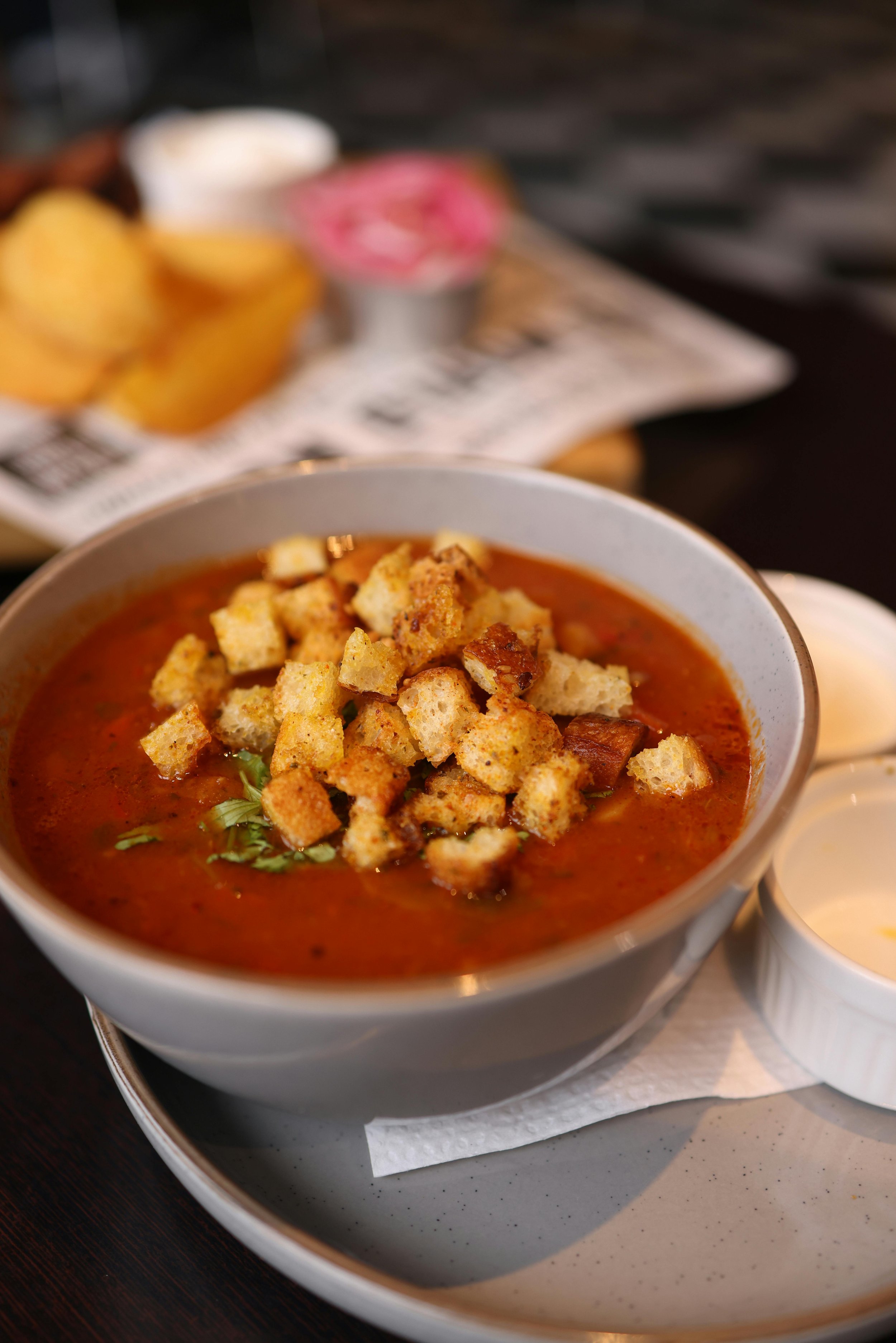 Bowl of tomato soup topped with croutons, served on a white plate with a side of creamy dressing.