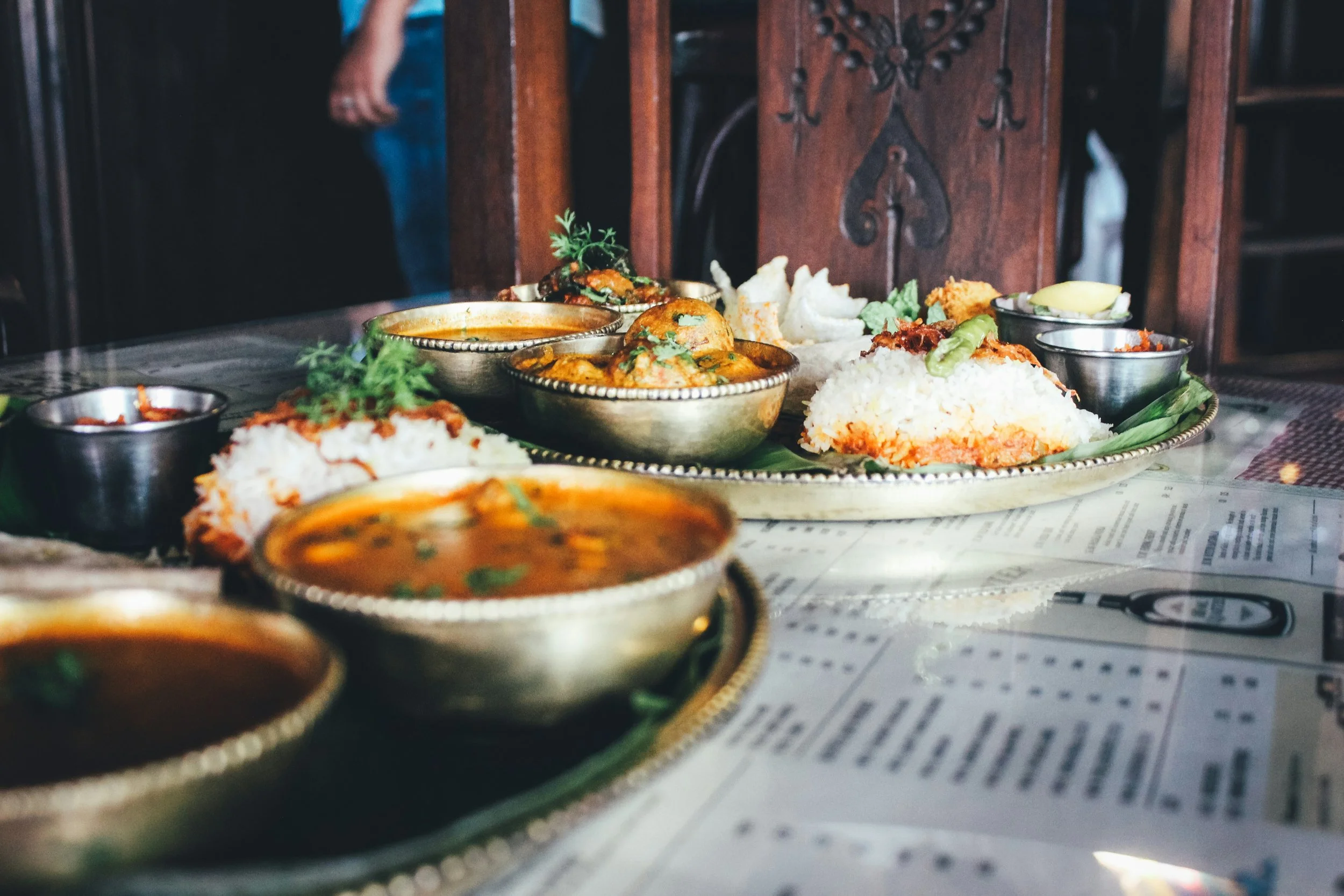 A table filled with traditional Indian dishes including bowls of curry, rice, and assorted condiments, set on a glass table with a mint leaf garnish, with a background of wooden furniture.