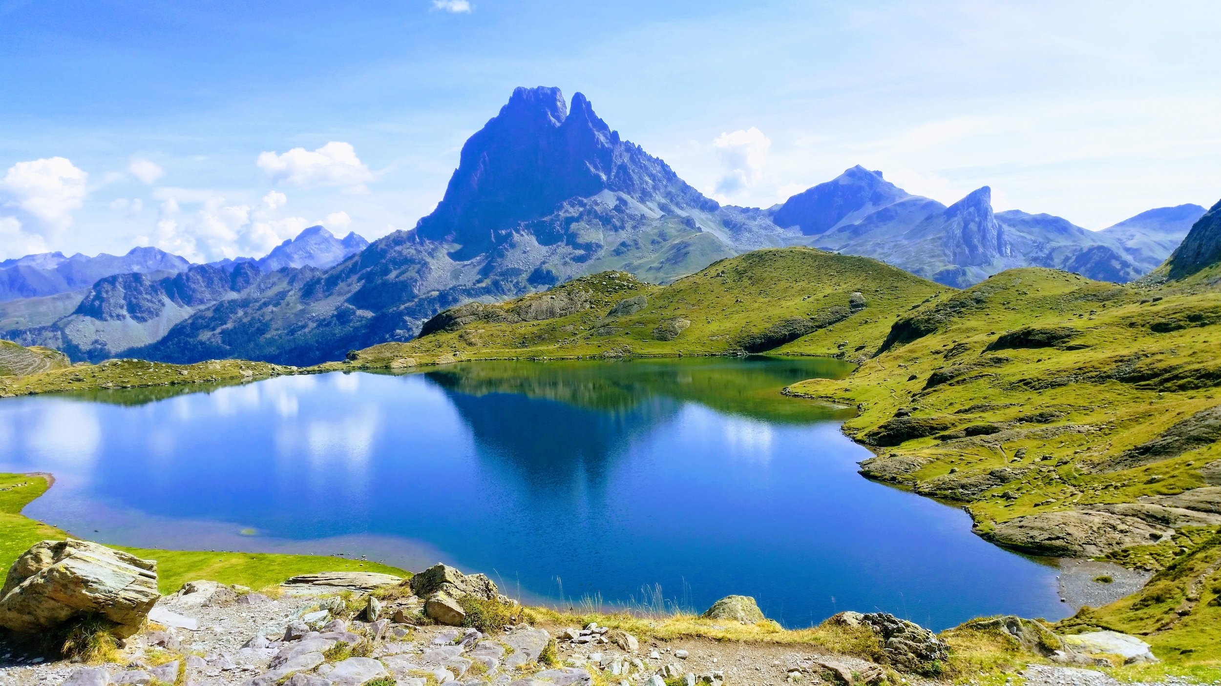 Scenic mountain landscape with a clear blue lake, green rolling hills, and towering mountain peaks under a partly cloudy sky.