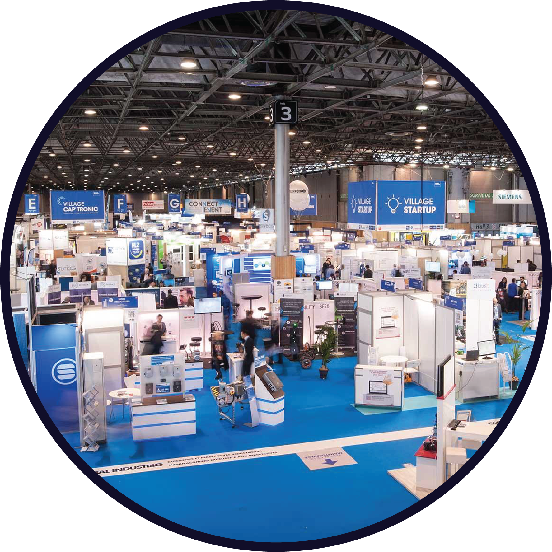 Salle d'exposition industrielle avec plusieurs stands, panneaux d'information bleus et blanc, et visiteurs.