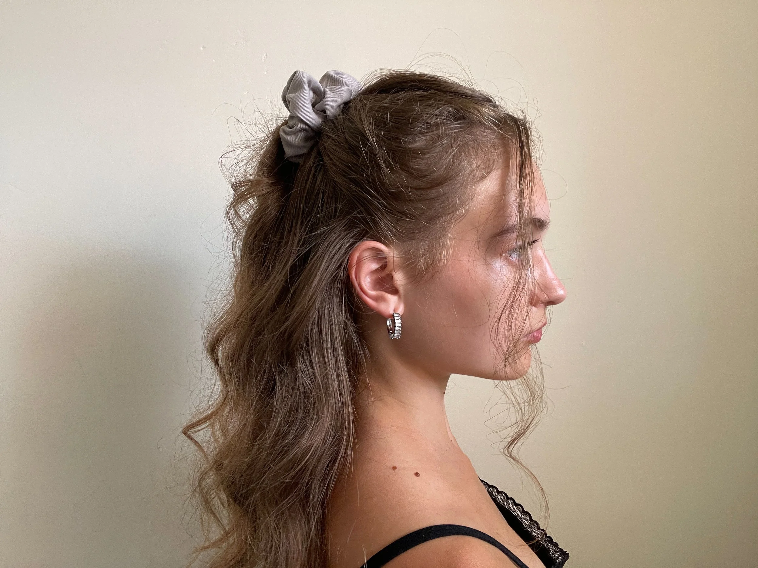 Profile of a woman with wavy brown hair, a gray scrunchie, silver hoop earrings, and a black tank top standing against a plain beige wall.