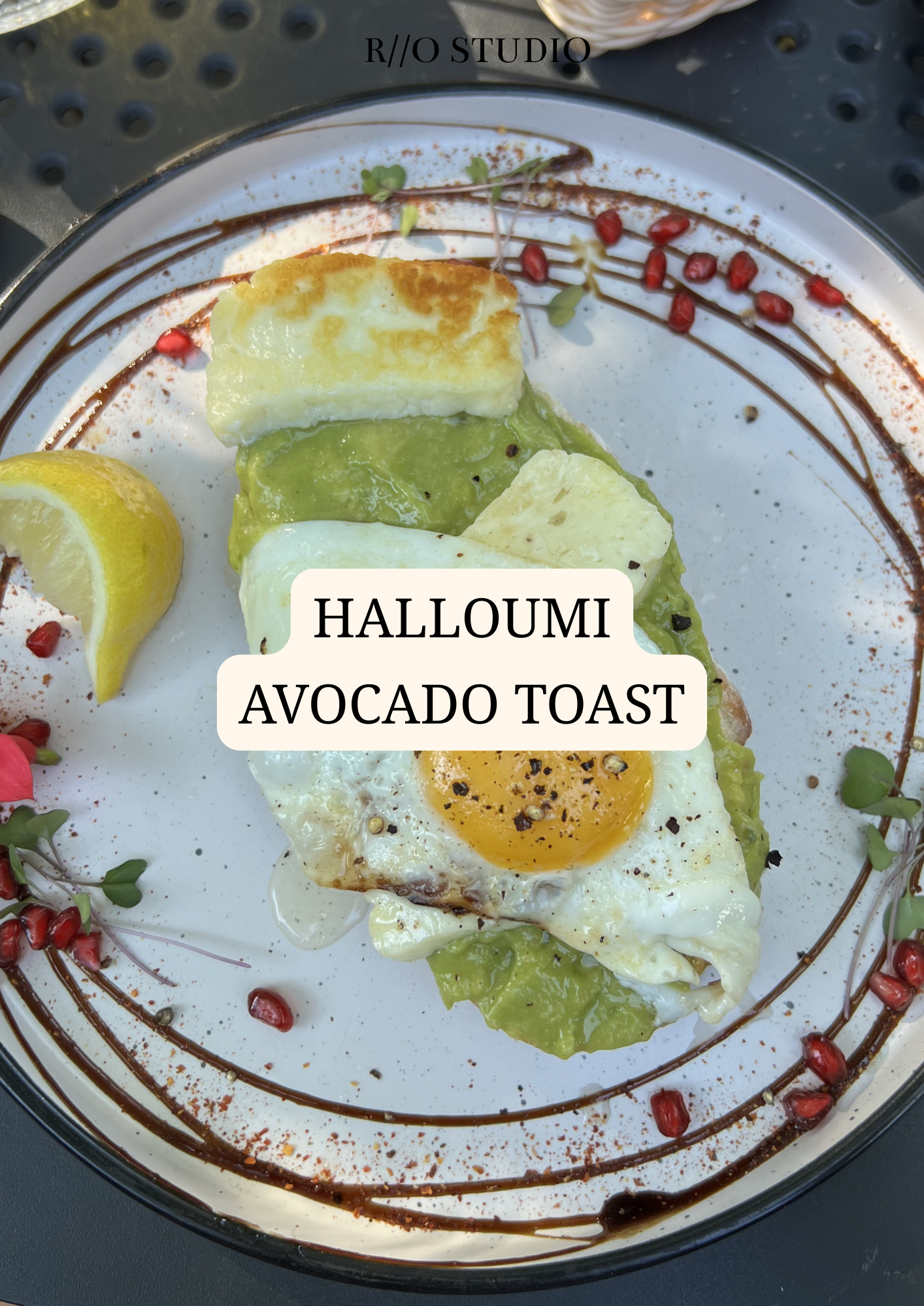 Plate with avocado toast topped with a fried egg, lemon wedge, garnished with pomegranate seeds, microgreens, and drizzled with balsamic glaze.