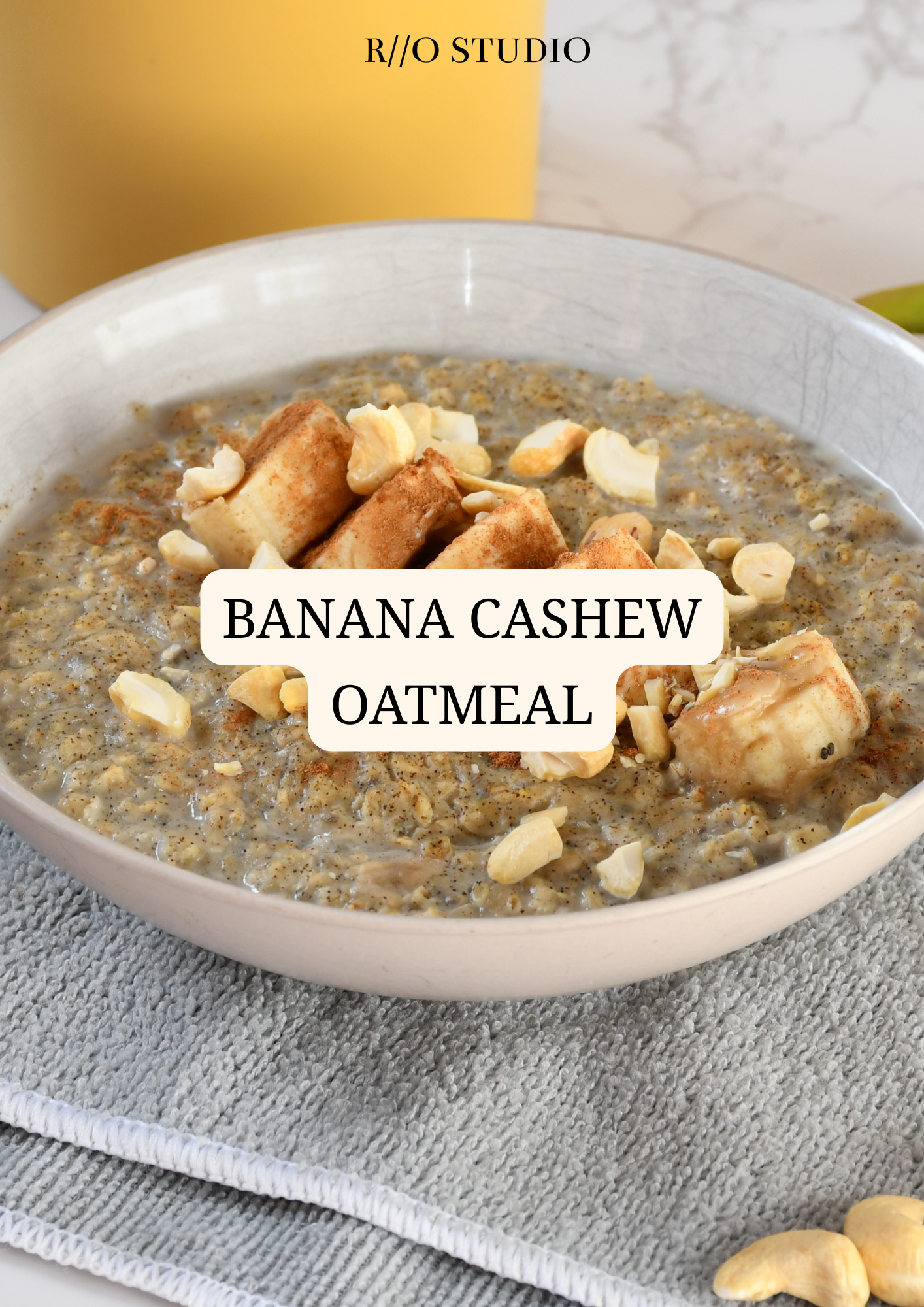 A bowl of banana cashew oatmeal topped with cashew pieces and cinnamon, with a yellow container and marble-patterned background in the background.