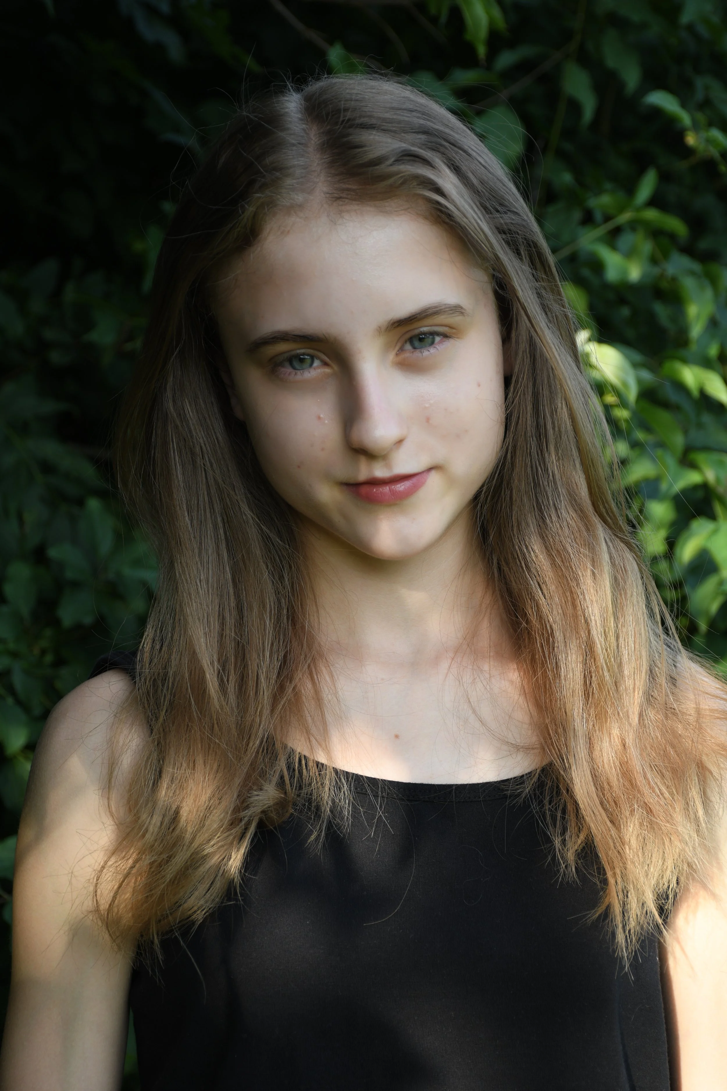 A young woman with long brown hair and blue eyes standing outdoors in front of green foliage.