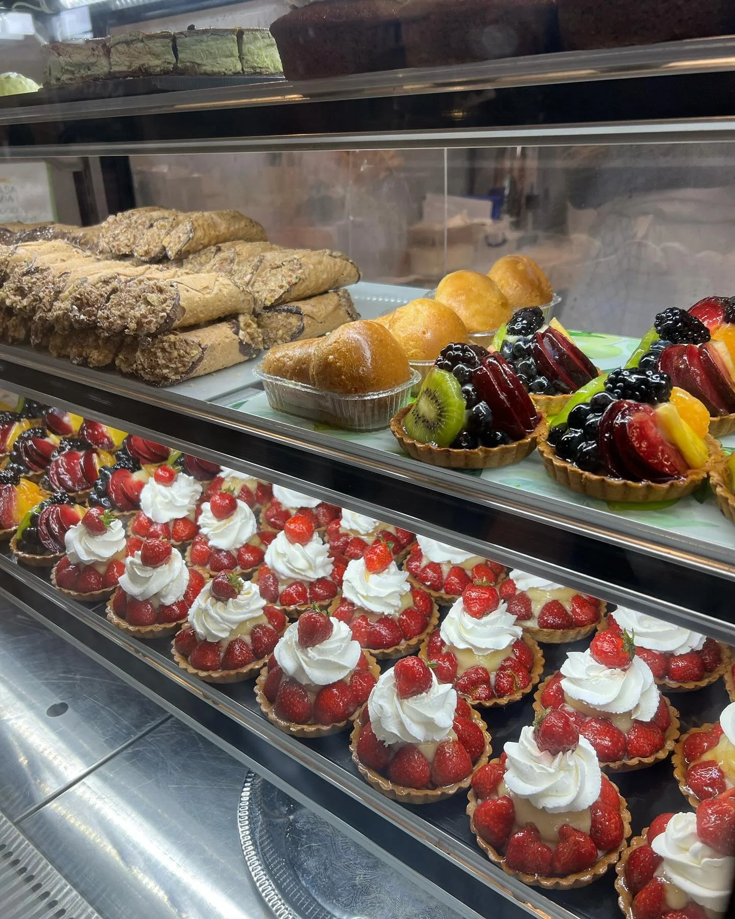 Display case with assorted baked goods including fruit tartlets topped with various berries and whipped cream, flaky pastries, and cookies.