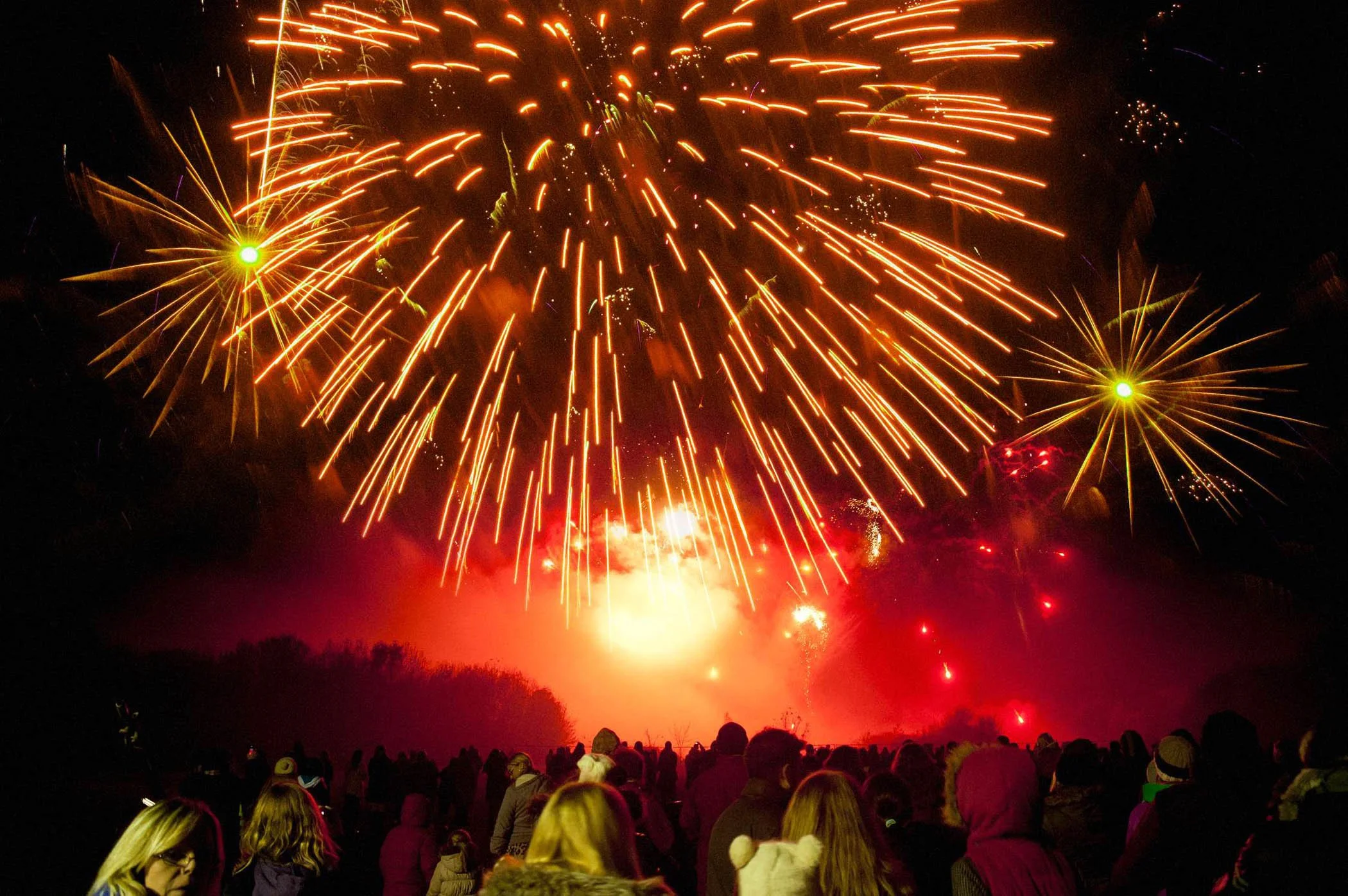 Nighttime scene with large, bright fireworks exploding in the sky, illuminating the crowd of people watching below. The fireworks are primarily orange, yellow, and red, creating a vibrant display at Kaboom in Rawcliffe Country Park, York