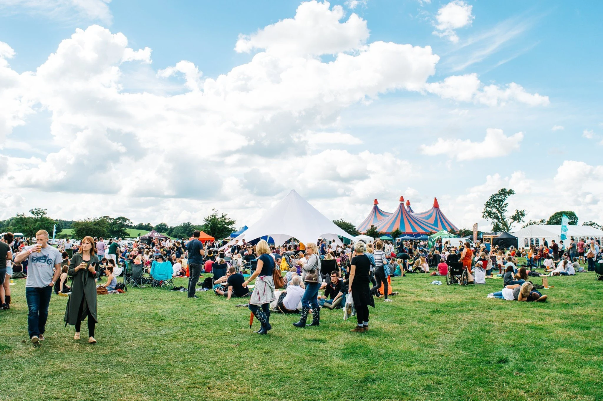 People attending an outdoor event or festival on a grassy field with tents and a circus tent in the background, under a partly cloudy sky.