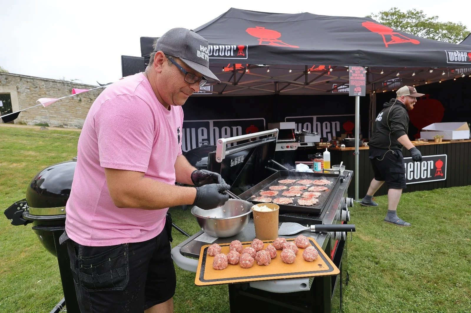 Man in pink shirt and black gloves preparing hamburgers on a grill at The Whole Hog BBQ competition, with another person in the background cooking at a food stall under a black tent.