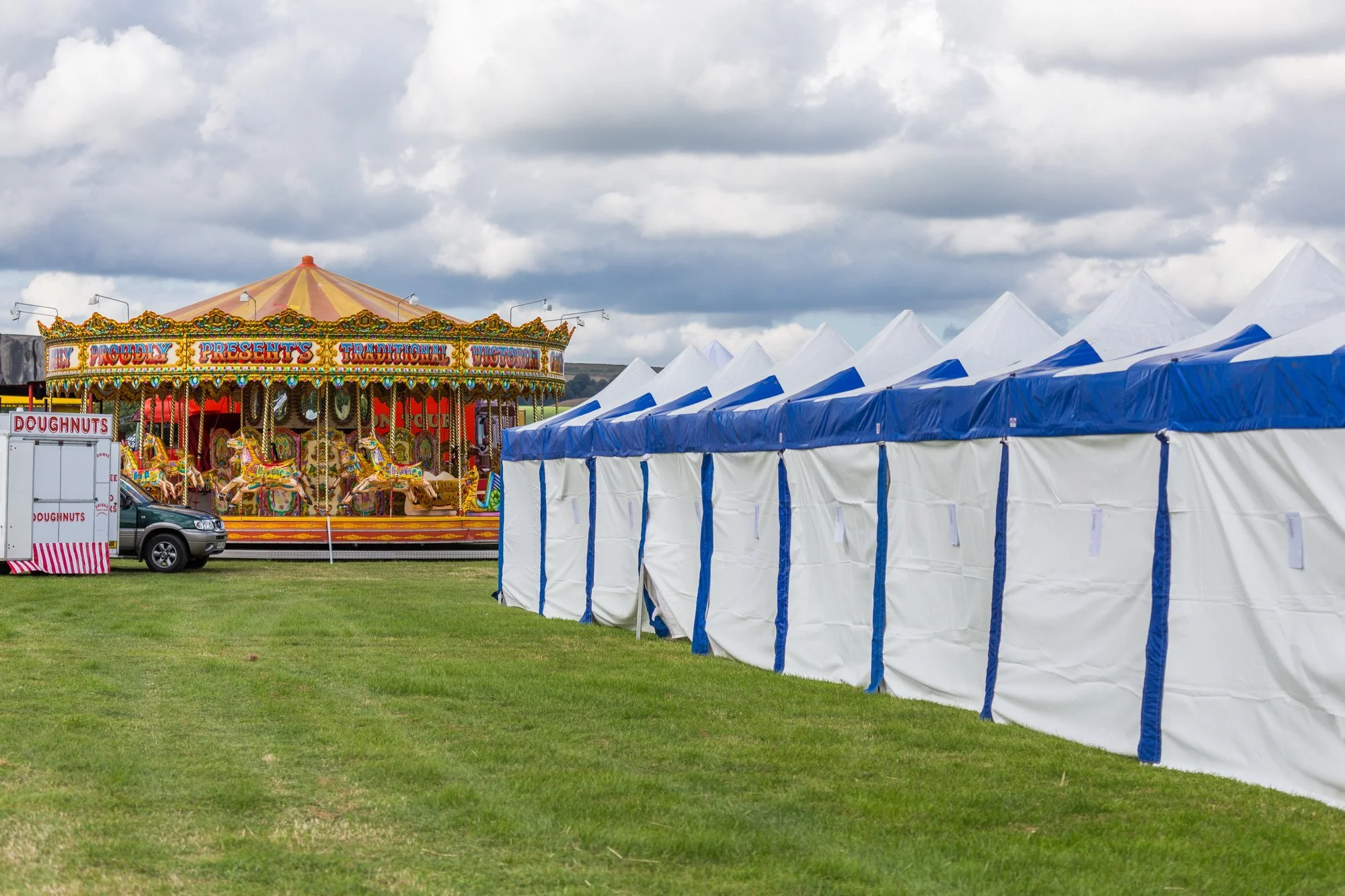 Empty market stall tents with a carousel ride in the background and a doughnut stand nearby on a cloudy day.