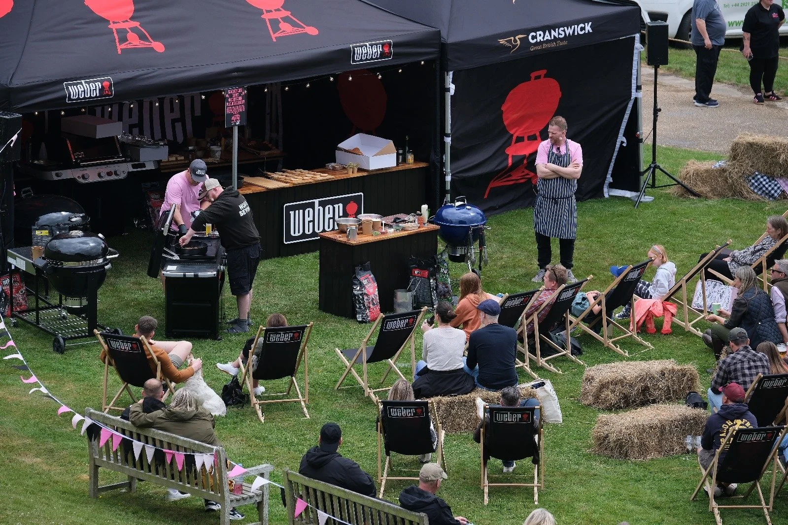 The Whole Hog BBQ competition with people sitting on deck chairs and hay bales, watching a barbecue demonstration on a stage with black tents and banners, including Weber and Cranswick logos.