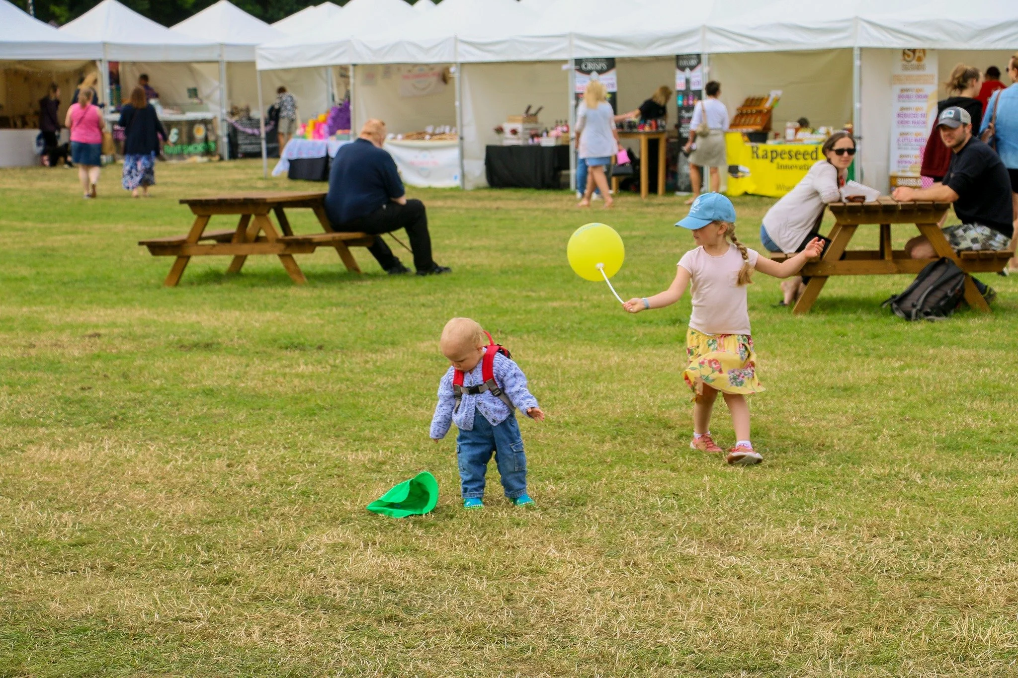 Two young children playing on the grass at an outdoor fair or festival with tents and food stalls in the background.
