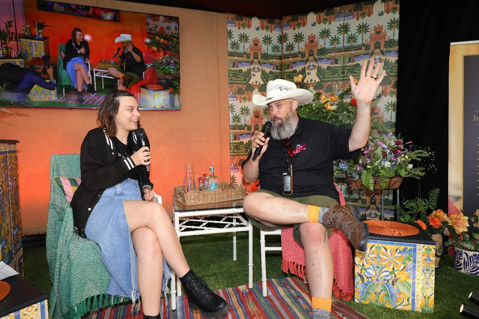 Two people sitting and talking with microphones, one woman and one man, in a colorful indoor setting with Mexican-themed decor and plants, a large screen behind them showing a panel discussion, both smiling and engaged in conversation.