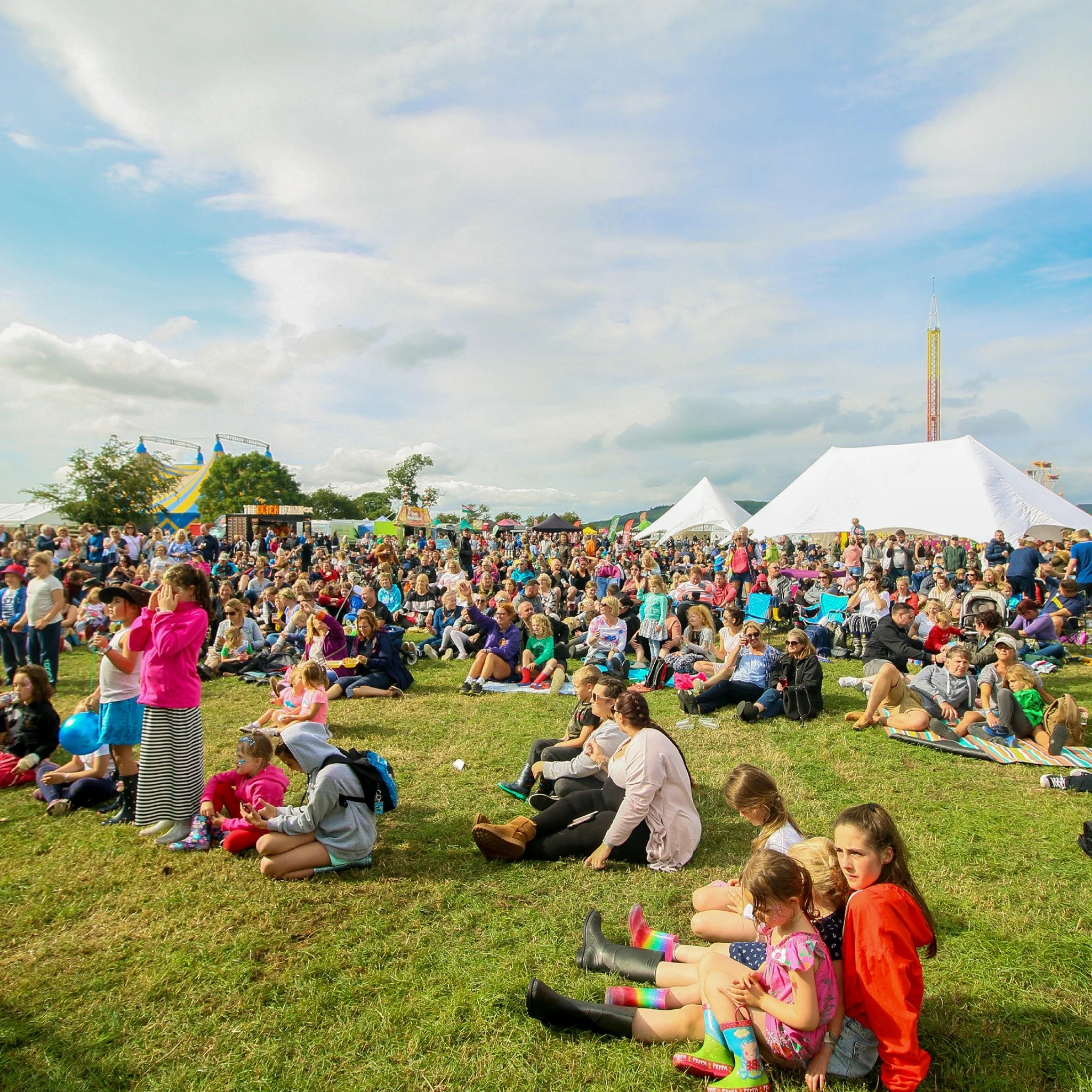 Crowd of people sitting and relaxing on grass at outdoor fair or festival with tents, rides, and cloudy sky in background.