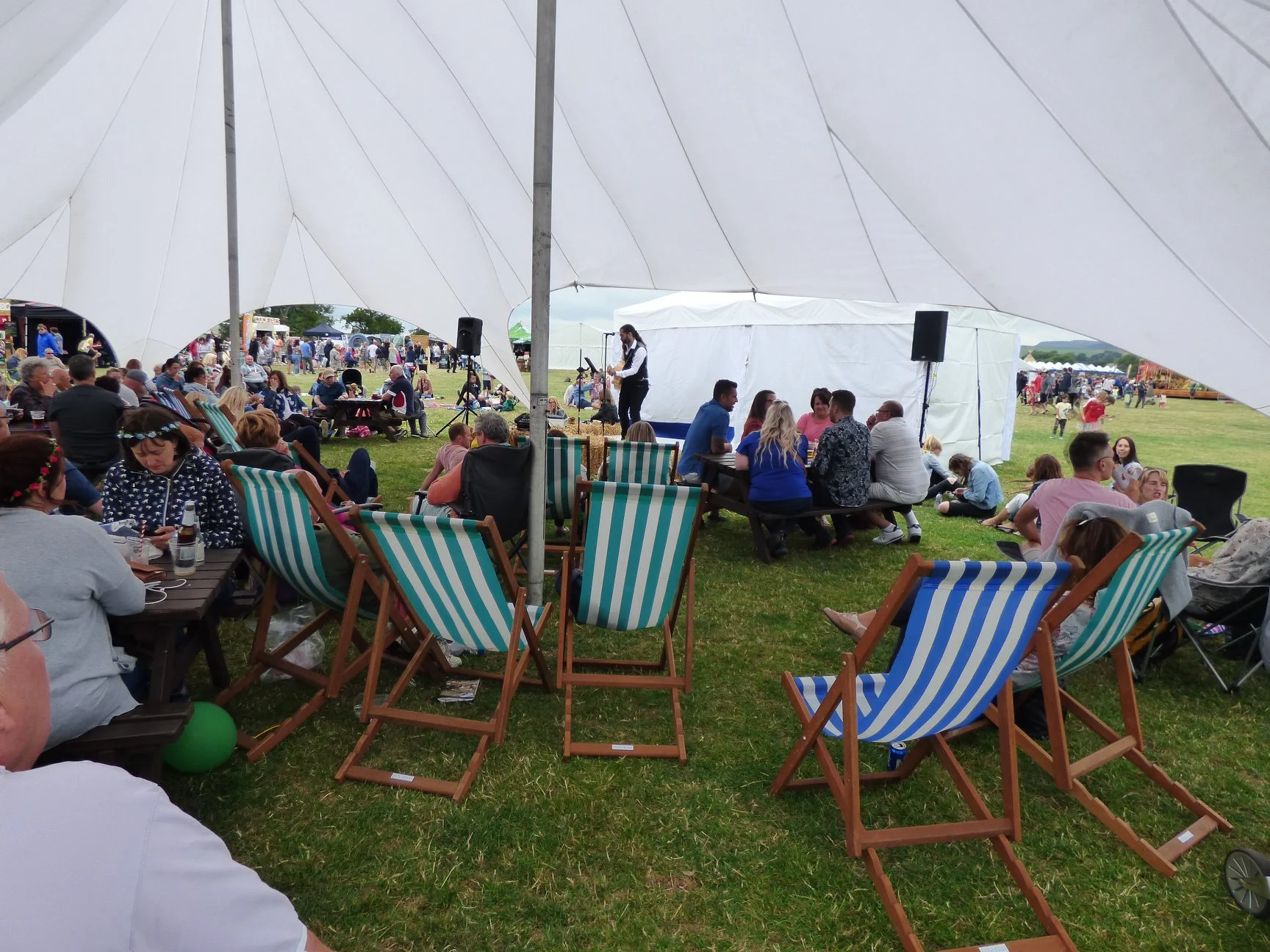 People gathering at an outdoor event under a large white tent with seating, a stage, and a musician playing guitar.