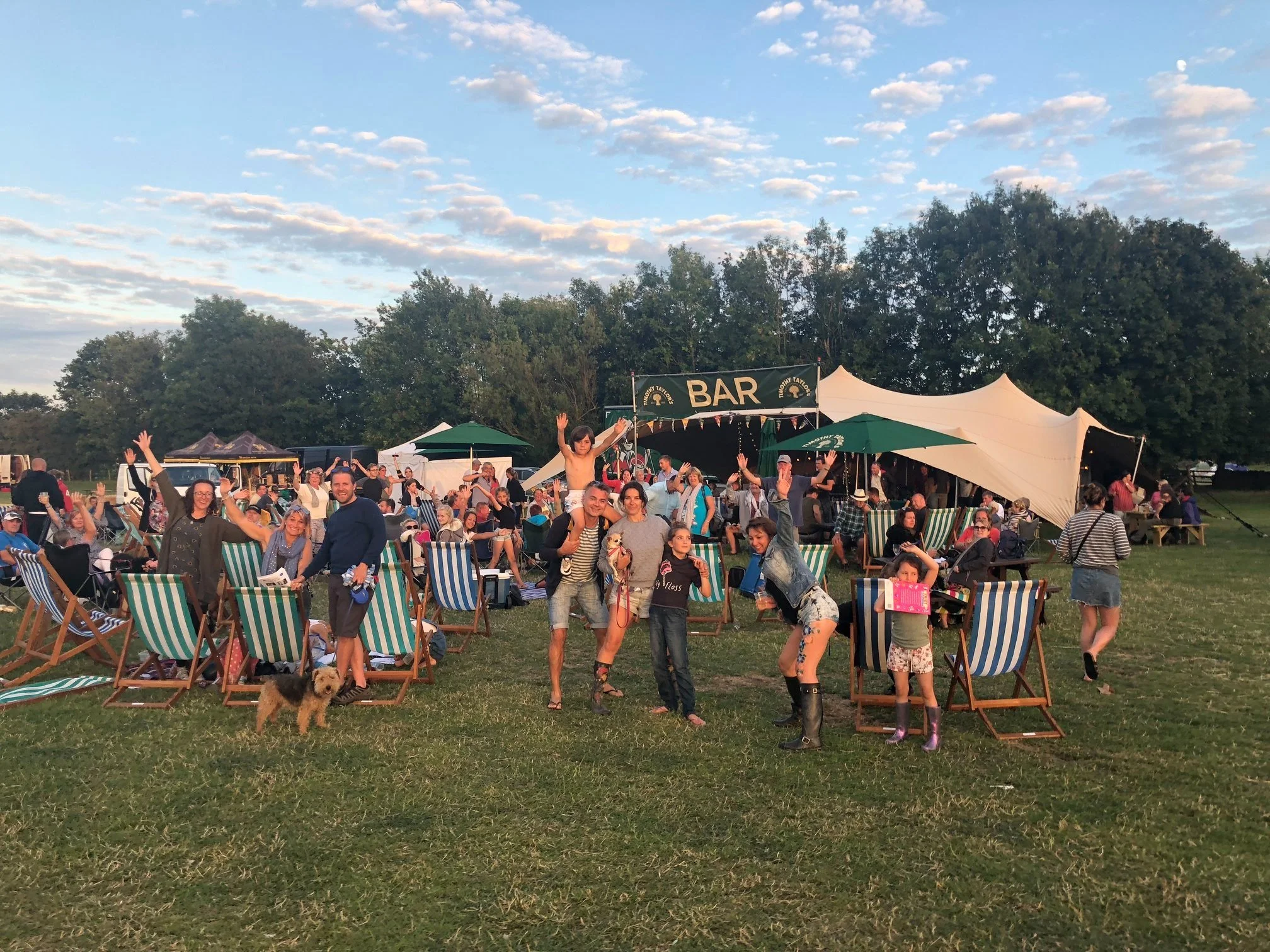 Outdoor gathering of people at a festival or fair with a bar and tents, some people sitting on striped chairs, some standing and waving, children playing, dogs and trees in the background under a blue sky with clouds.