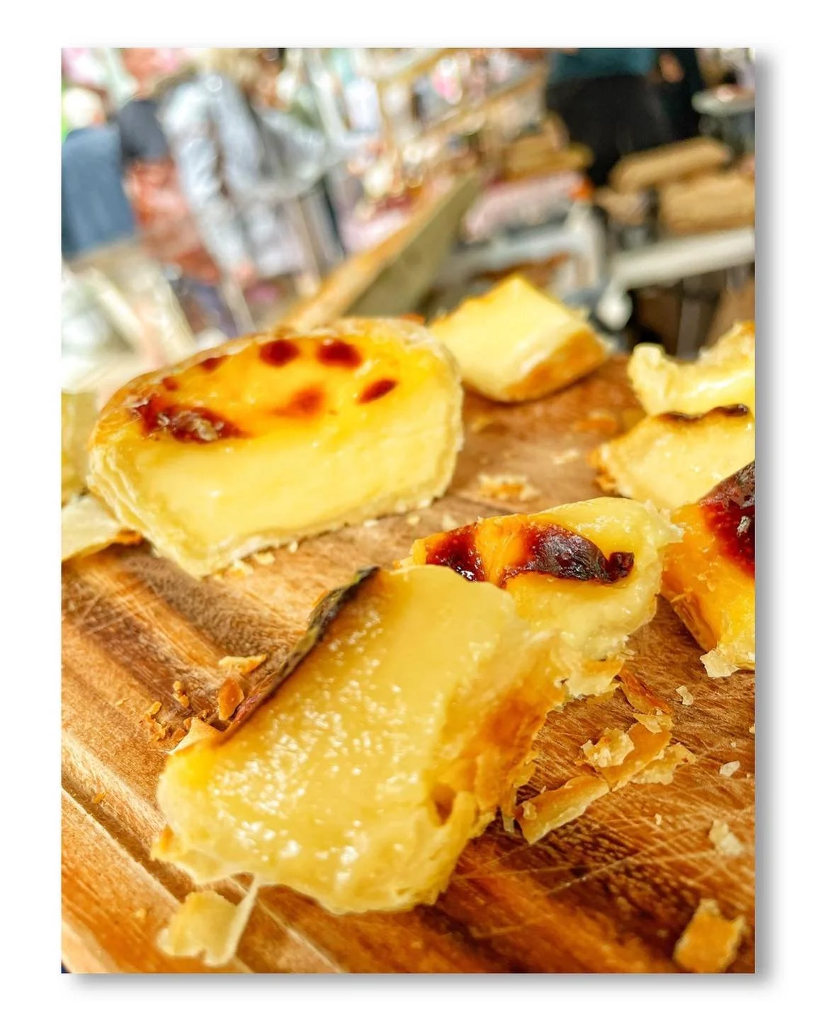 Close-up of cheesy, baked potato slices on a wooden cutting board with a blurred background of a market or food stall.