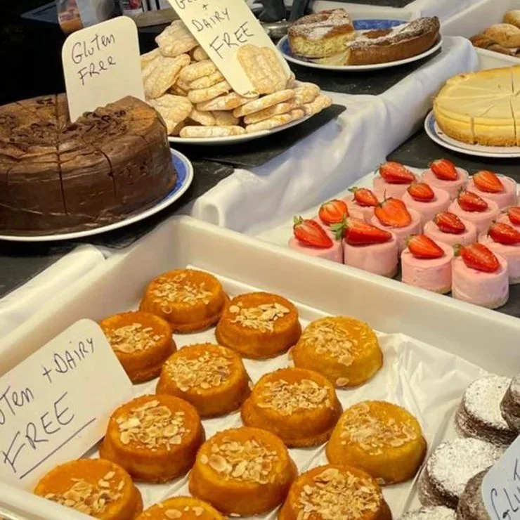 A variety of desserts including a chocolate cake, plate of cookies labeled gluten-free, lemon bars, strawberry-topped pink cakes, caramel donuts with almond slices, and chocolate donuts with powdered sugar.