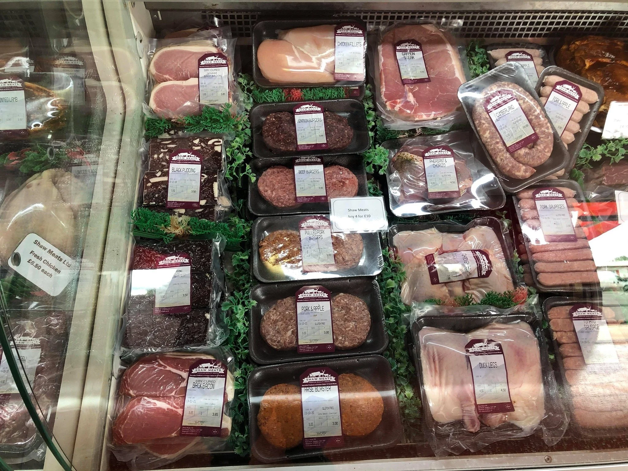 Display of various packaged meats including chicken fillets, steak, burgers, pork, bacon, and sausages in a refrigerated case at a butcher's market stall.