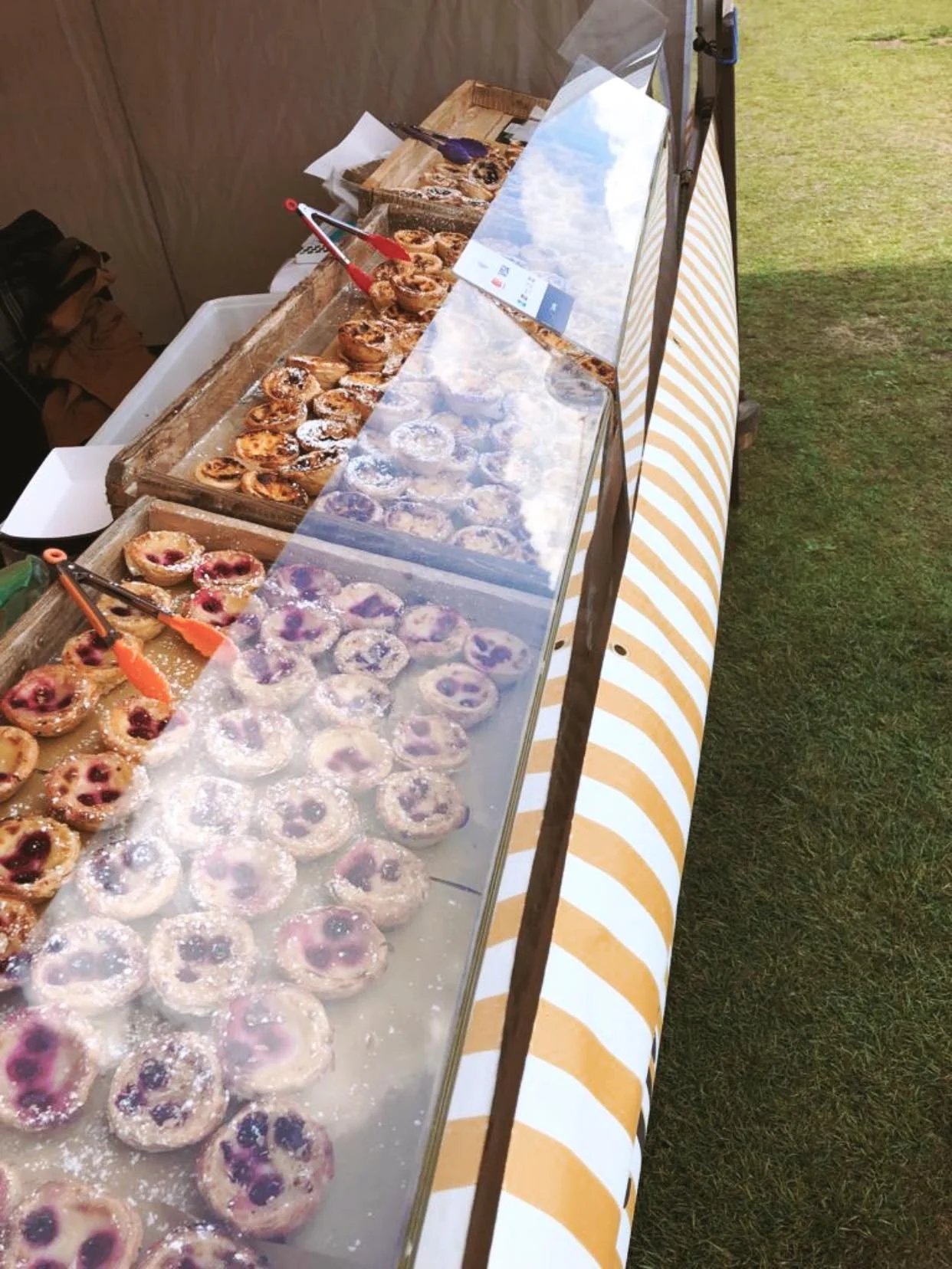 Display of various pastries on a tray at an outdoor event, some covered with transparent plastic, with tongs available for serving.