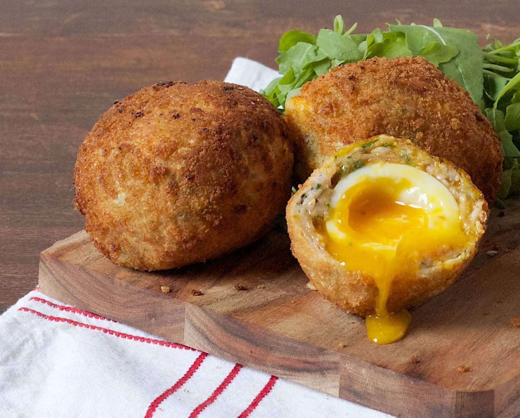 Three golden-brown Scotch eggs on a wooden board, one cut in half revealing a soft-boiled egg inside, garnished with greens.