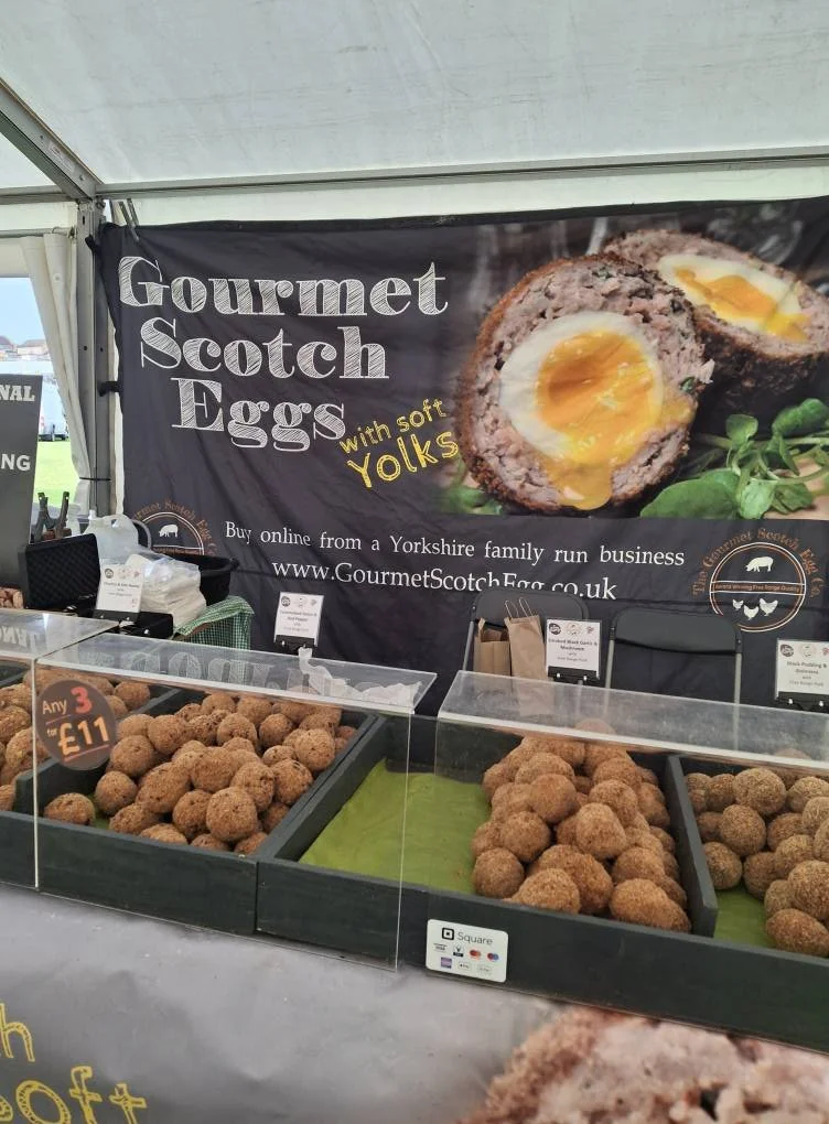 Display of gourmet Scotch eggs with soft yolks, sold in a market stall with a banner advertising the product and a website link.