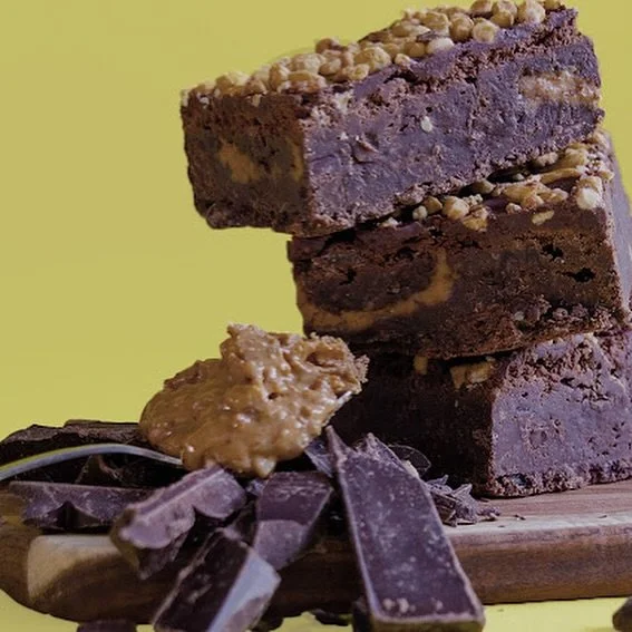 Stacked chocolate brownies with nuts on top, chocolate chunks, and peanut butter on a wooden board with a yellow background.