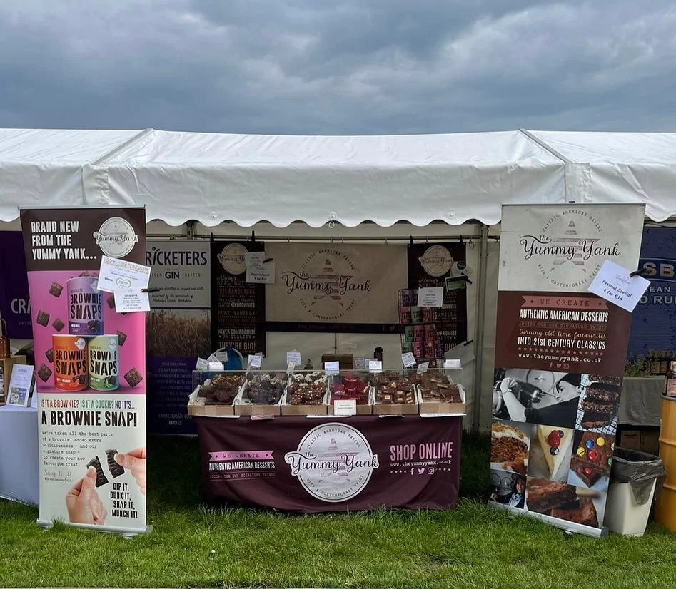 A market stall under a white tent displaying baked goods and desserts from Yummy Yank, with banners and signs promoting American-style desserts and brownies, on a grassy outdoor area with cloudy sky overhead.
