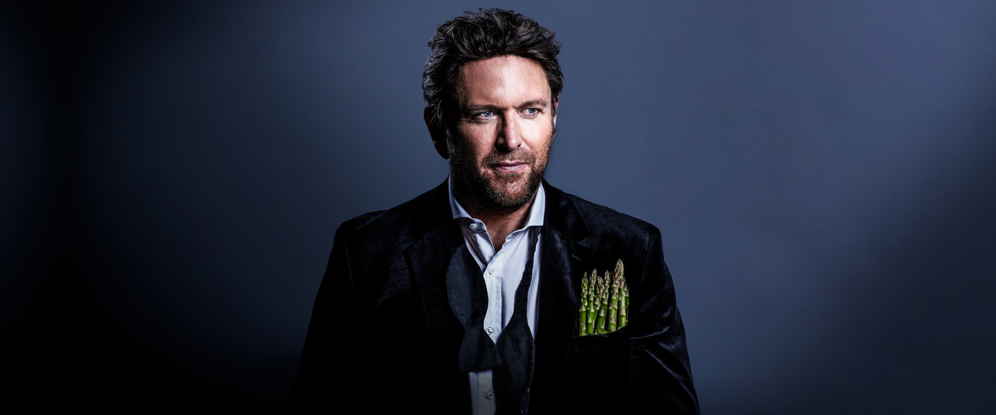 A photo of celebrity chef James Martin wearing a black velvet blazer with asparagus in his breast pocket, set against a dark gradient background.
