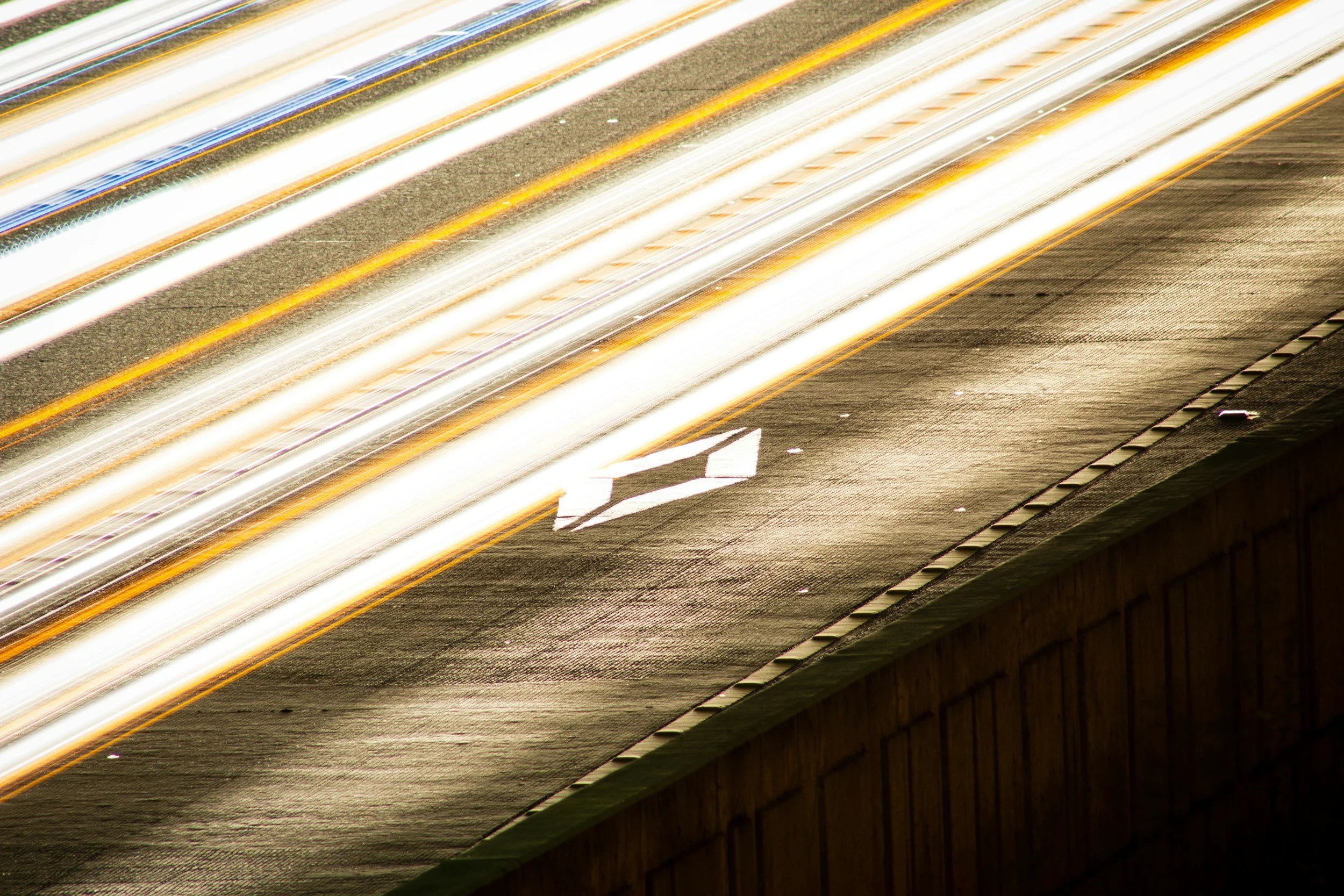 Light trails from moving vehicles on a highway at night, with lane markings and a street sign visible on the road.