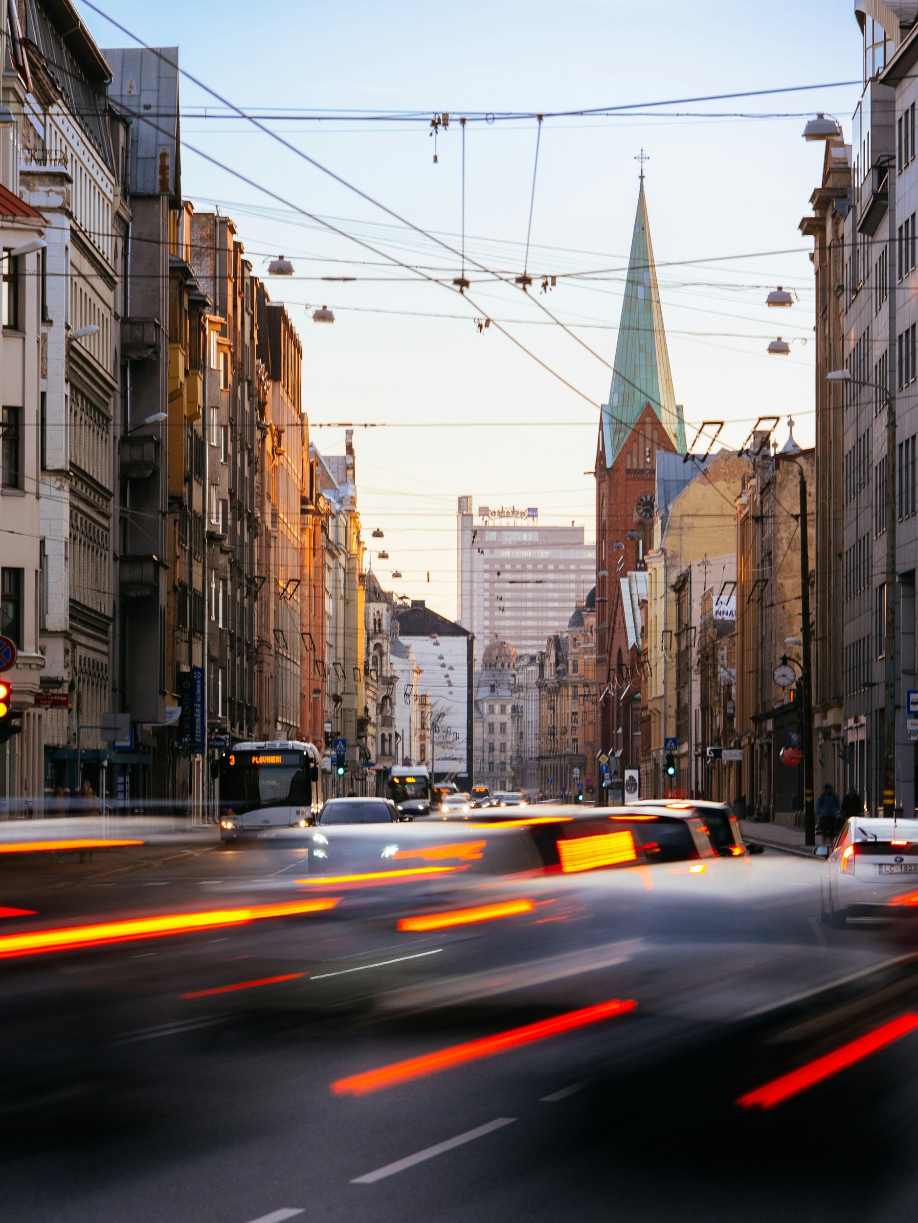 Traffic on a city street during sunset with moving cars, tall buildings, and a church with a green spire in the background.