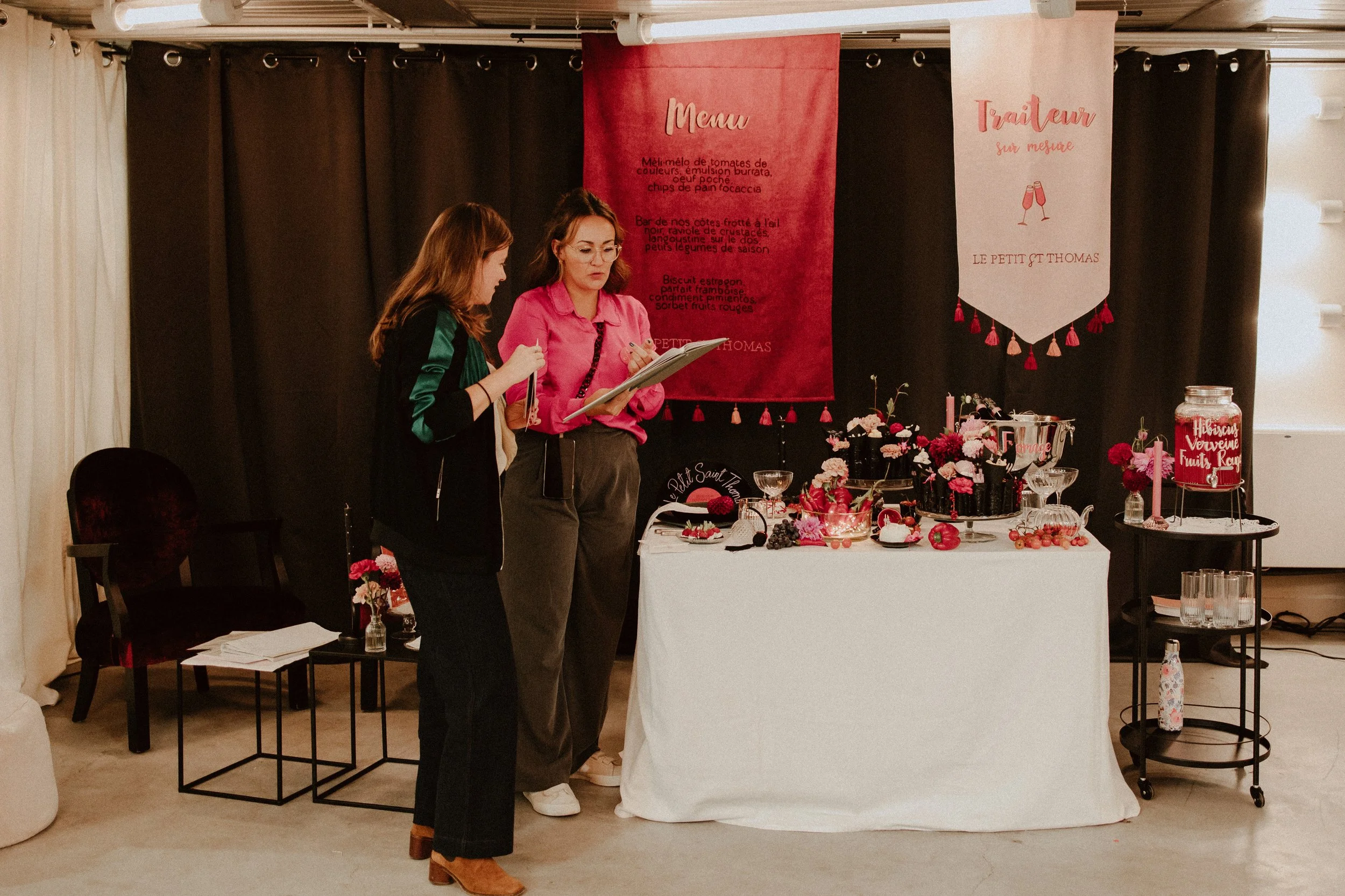 Deux femmes discutent près d'une table avec des desserts et des fleurs, dans une pièce décorée de bannières roses et rouges pour un événement ou un dîner thé.