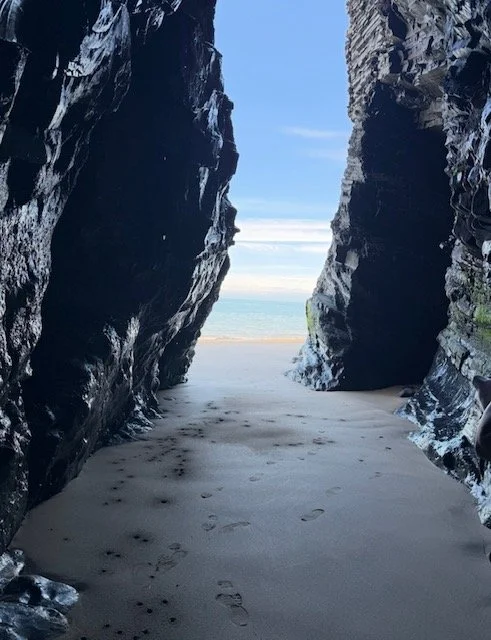 View of sandy beach seen through a narrow rocky passage between two cliffs with footprints in the sand, leading to the ocean in the distance.