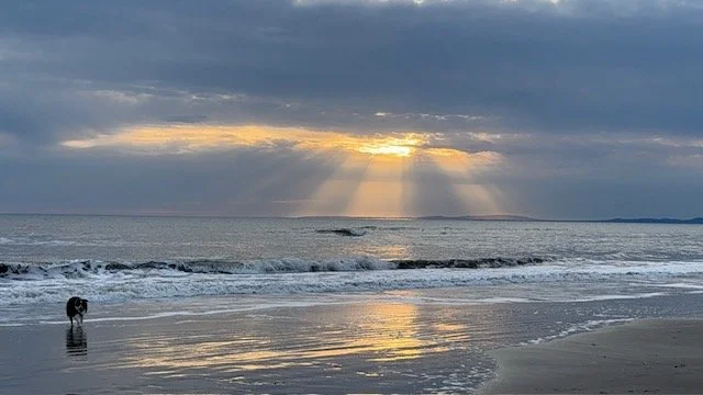 Sunset over the ocean with rays of sunlight breaking through clouds, waves crashing on a sandy beach, and a person walking along the shoreline.