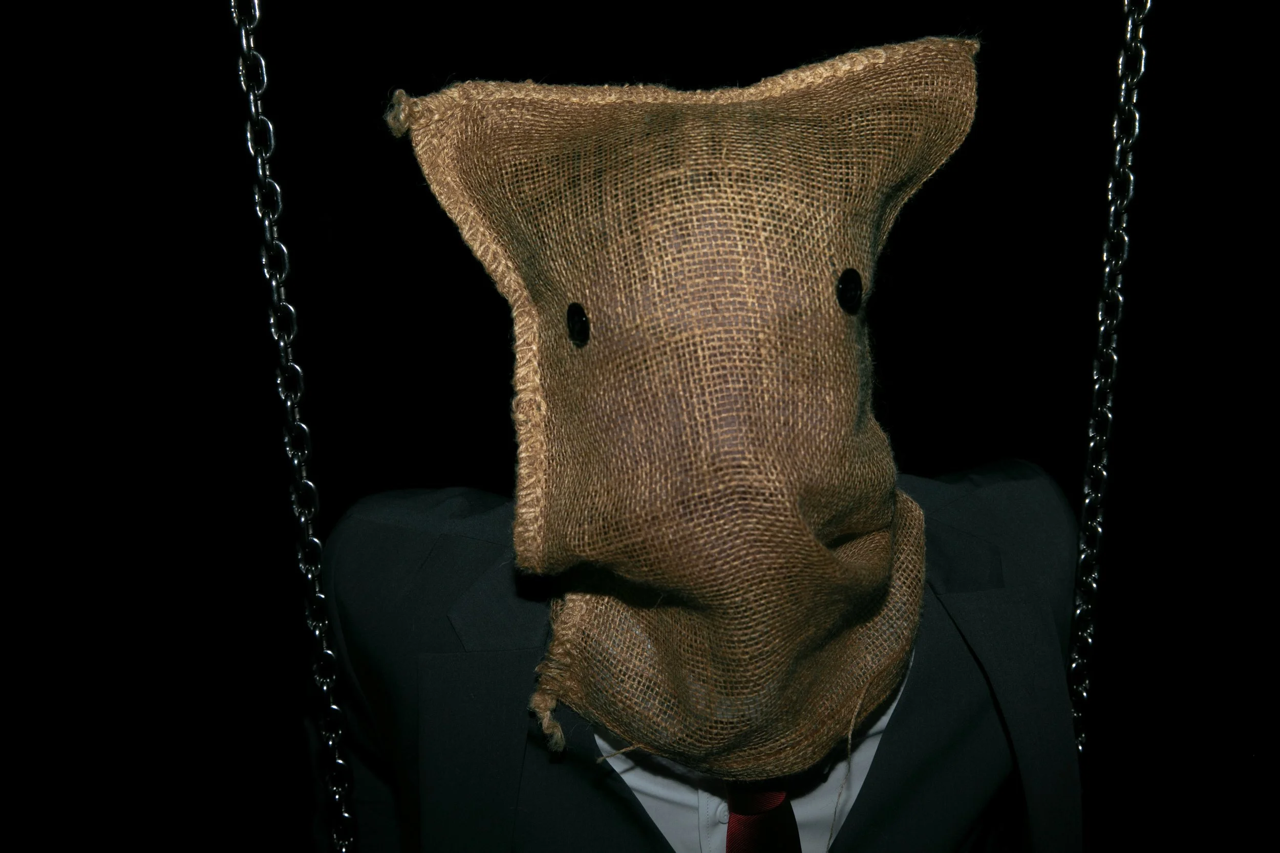Person wearing a burlap sack with eye holes over their head, dressed in a suit and tie, sitting on a chain-linked chair against a black background.