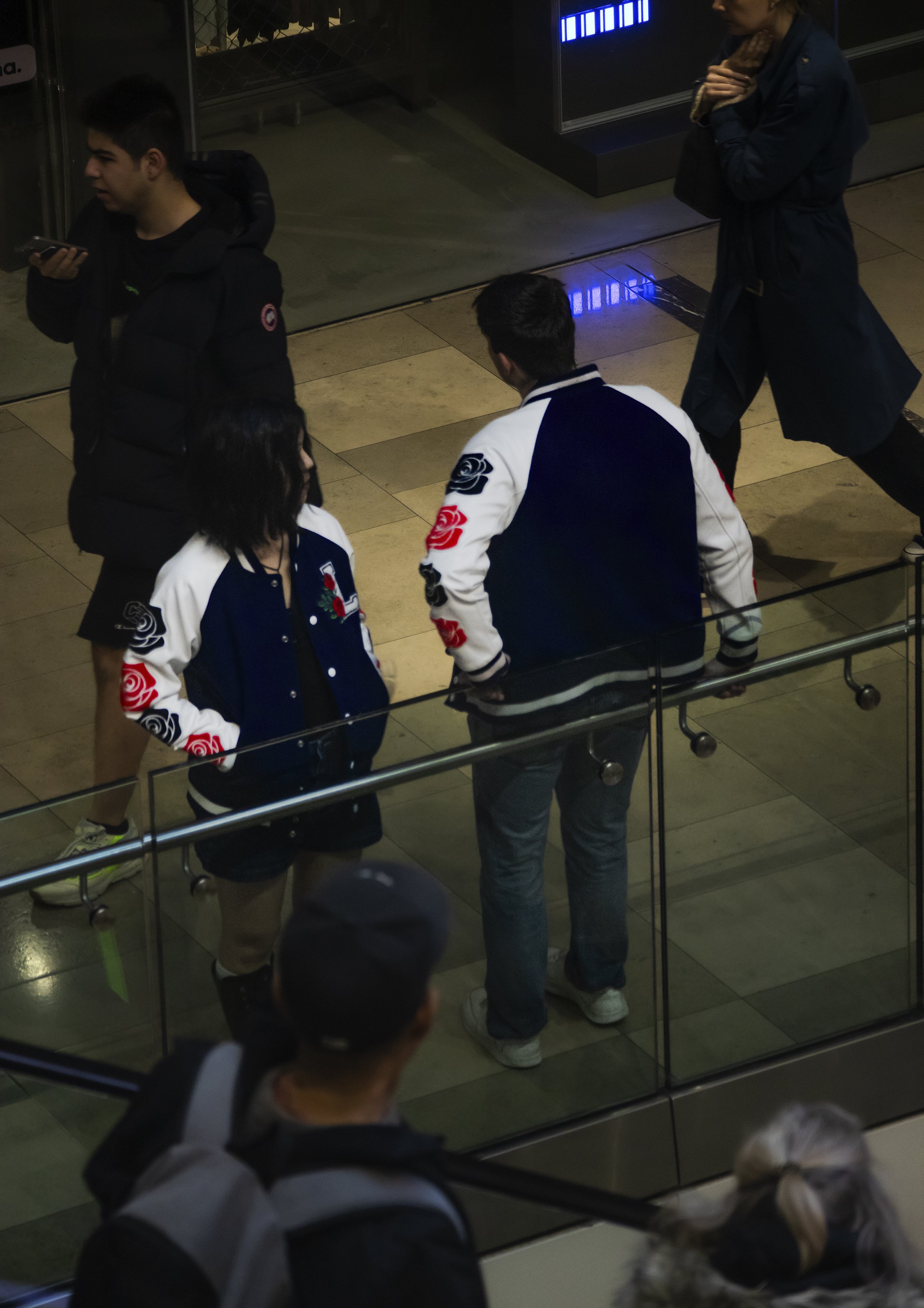 Two young adults, a man and a woman, standing and talking inside a shopping mall near a glass barrier, wearing varsity jackets with floral designs on the sleeves. Two other people, a man and a woman, are walking by in the background.