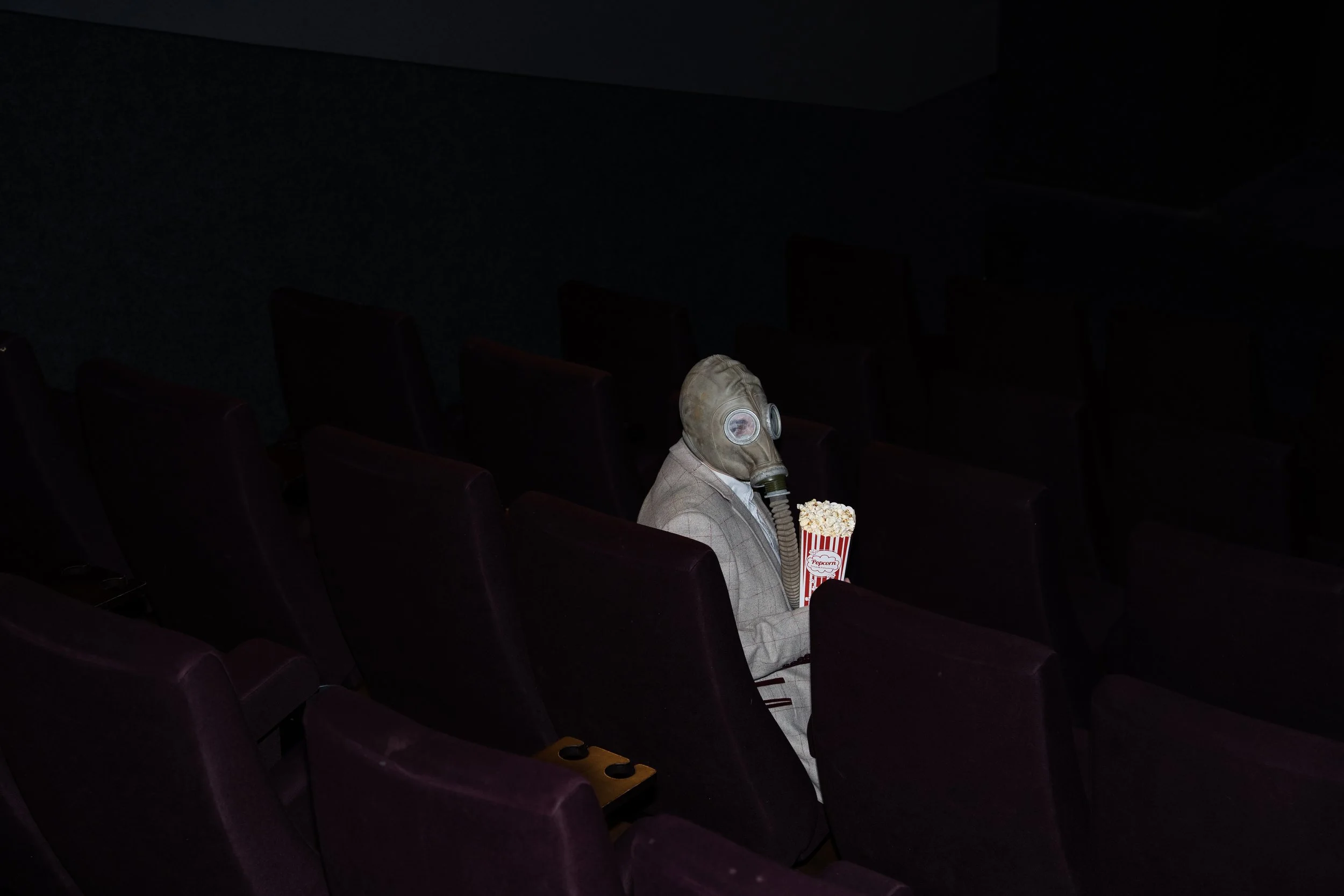 A person wearing a gas mask and a suit sitting alone in an empty movie theater, holding a bucket of popcorn.