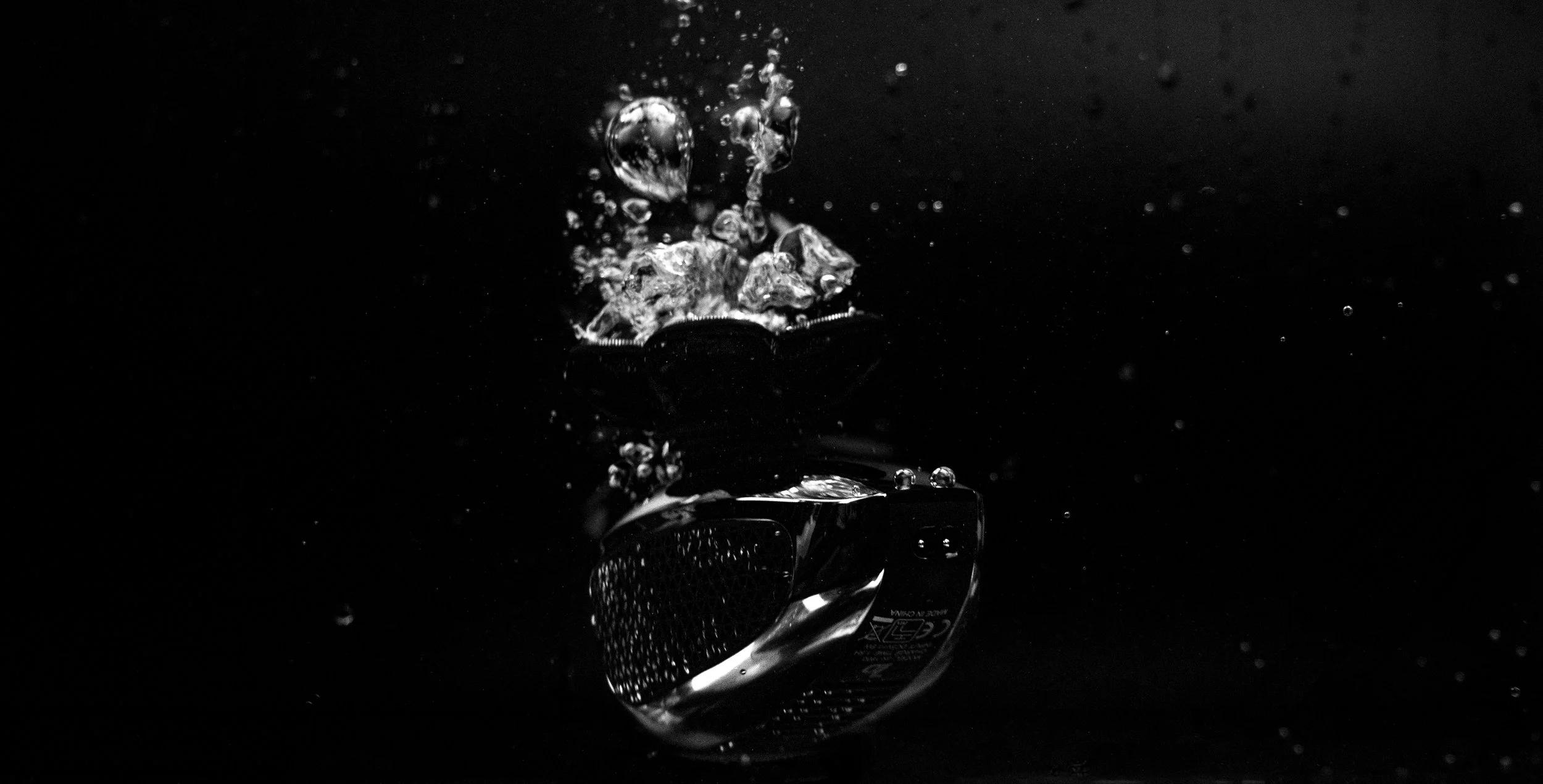 A close-up of a black object with a textured surface, submerged underwater with bubbles rising above it, against a dark background.