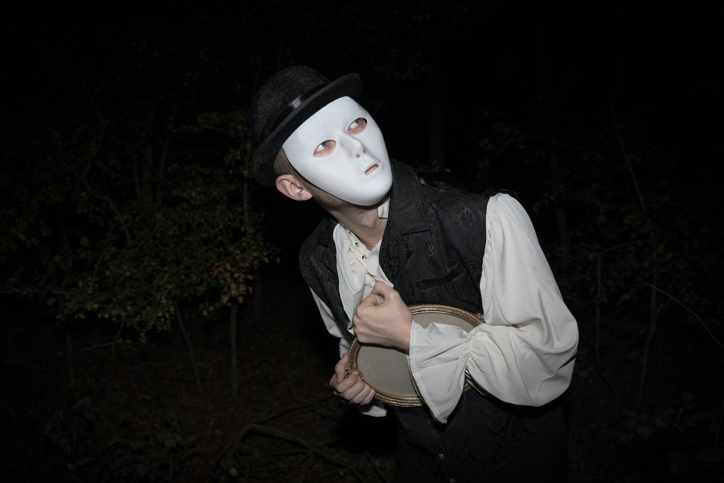 Person wearing a white mask, black hat, and vintage clothing, holding a small mirror, standing outdoors at night in a dark wooded area.