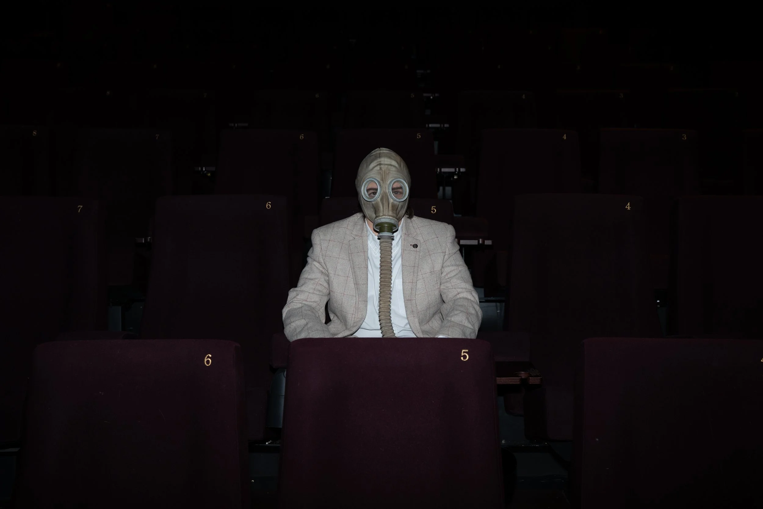 Person sitting alone in a dark theater wearing a gas mask and a beige plaid blazer.